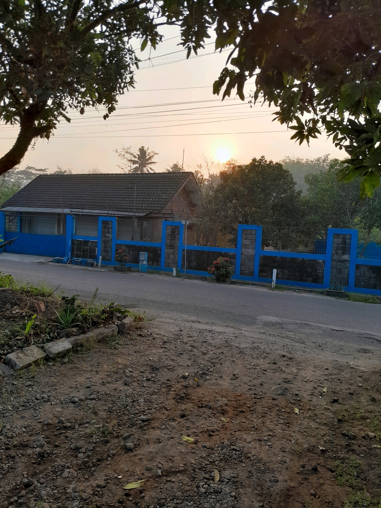 The image depicts a scene in Glanggang, Indonesia, at sunrise. A building with a dark tiled roof and blue accents stands beside a street. A fence with blue decorative elements and dark stone blocks separates the property from the road. In the foreground, the ground is covered with small stones and dirt, with some vegetation growing along the roadside. Trees with green leaves frame the top of the image, and the rising sun is visible in the distance, casting a warm glow on the sky and the surrounding landscape. The environment appears to be rural or semi-rural, with a peaceful and serene atmosphere. There are no people visible in the image.