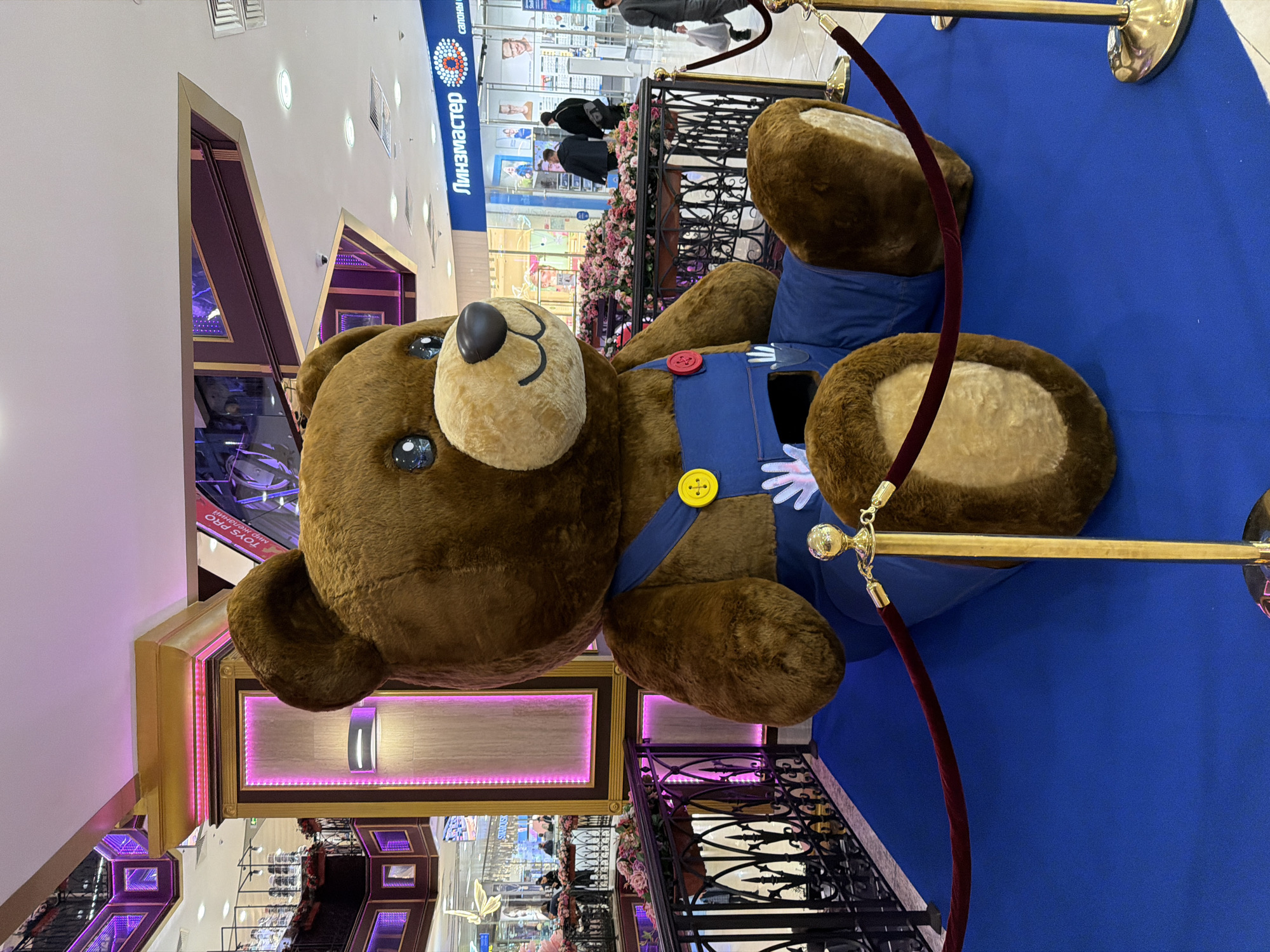 A large brown teddy bear wearing a blue outfit with yellow and red buttons lies on a blue surface, surrounded by gold stanchions with velvet ropes. The bear is positioned at the top of a staircase with decorative black metal railings adorned with floral arrangements. Behind the bear and railing is a shopping mall interior with visible storefronts, including one with the name "Линзмастер" displayed in Russian. The mall features multiple levels with shops and walkways. The background suggests a commercial setting, possibly a shopping center or mall. The location is Moscow, Russia, based on the signage in Russian.