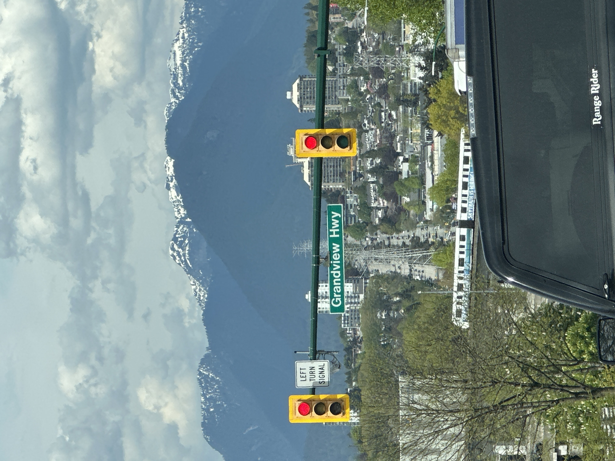 The image presents a daytime scene from an urban environment, with significant natural features in the background. The perspective is from inside a vehicle, evidenced by the dark frame on the right side of the image, where the text "Range Rider" is visible.
In the immediate foreground, two traffic light assemblies are prominent. The main traffic light, which is yellow-framed, displays a red signal for straight-ahead traffic. Below this, a smaller white sign clearly states "LEFT TURN SIGNAL," also indicating a red signal. A green street sign attached to the traffic light pole reads "Grandview Hwy."
Beyond the street infrastructure, the midground features a cityscape with numerous buildings and abundant green trees, suggesting a verdant area. Power lines crisscross the urban landscape. An elevated rapid transit train, specifically a SkyTrain, is seen moving along its tracks in the mid-distance; the text "SKYTRAIN IS HIRING!" is faintly discernible on its side.
The background is dominated by a range of majestic mountains, their peaks and upper slopes covered in snow, indicating a significant elevation. The sky above is a bright blue with scattered white, cumulus clouds. The lighting suggests it is a clear day, likely late morning or afternoon.
The snow-capped mountains and the presence of a SkyTrain, along with the "Grandview Hwy" sign, strongly suggest the location is Vancouver, Canada, aligning with the provided context. The overall impression is a bustling urban area set against a dramatic natural backdrop.