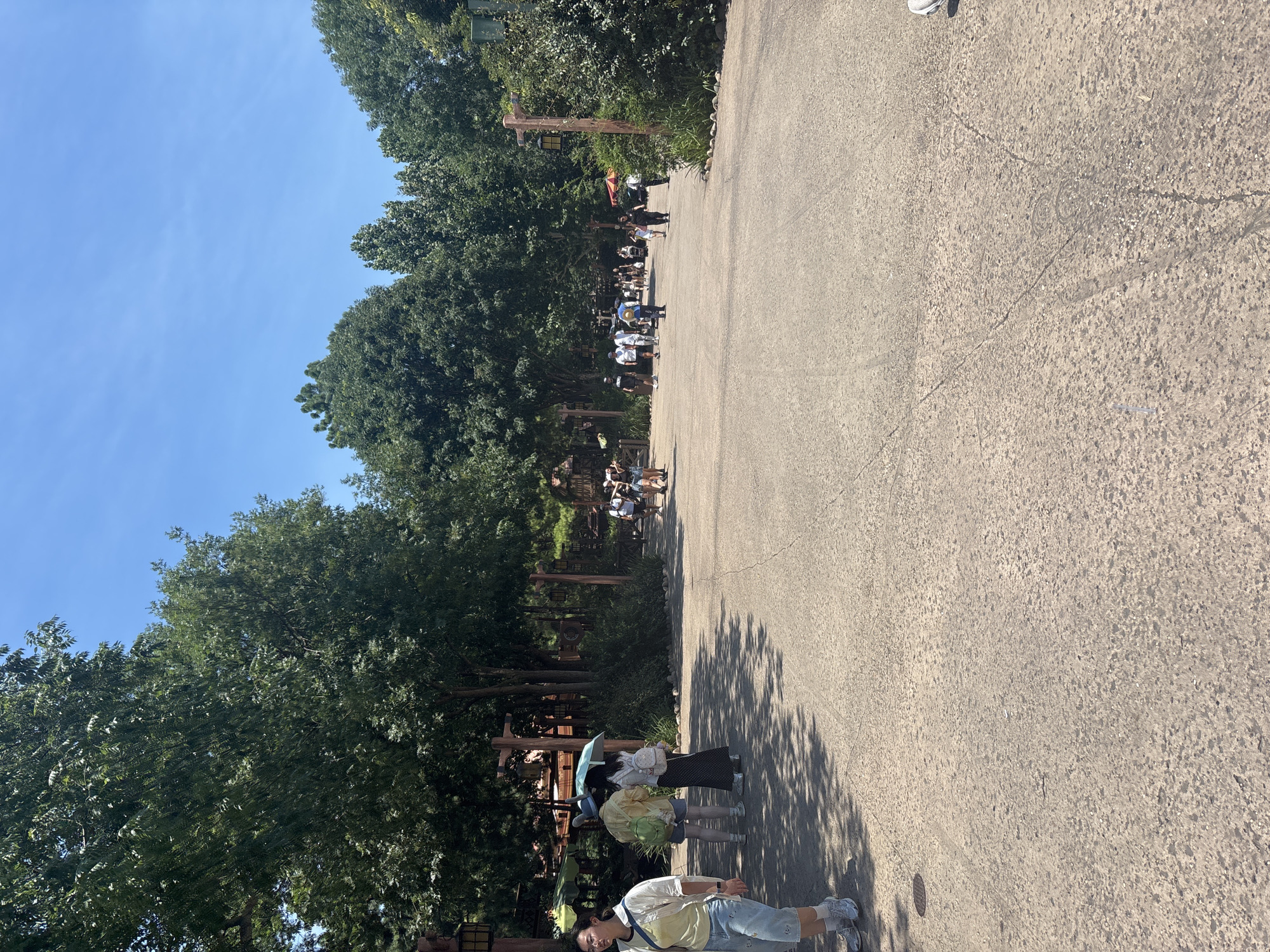 The image depicts a paved pathway through a park-like setting with lush green trees lining both sides. The sky is clear blue. Several people are visible walking along the pathway. In the foreground, a woman wearing a light-colored shirt and blue shorts is walking towards the left. To her right, a couple stands under a light blue umbrella, with one person wearing a yellow shirt and the other a white top. Further down the path, a group of people are seen walking in various directions. The overall impression is that of a sunny day in a public outdoor space. The location is identified as Pudong, China, which suggests an urban park or recreational area within that district. The architecture of the lamp posts and fences hints at a theme park or a cultural attraction.