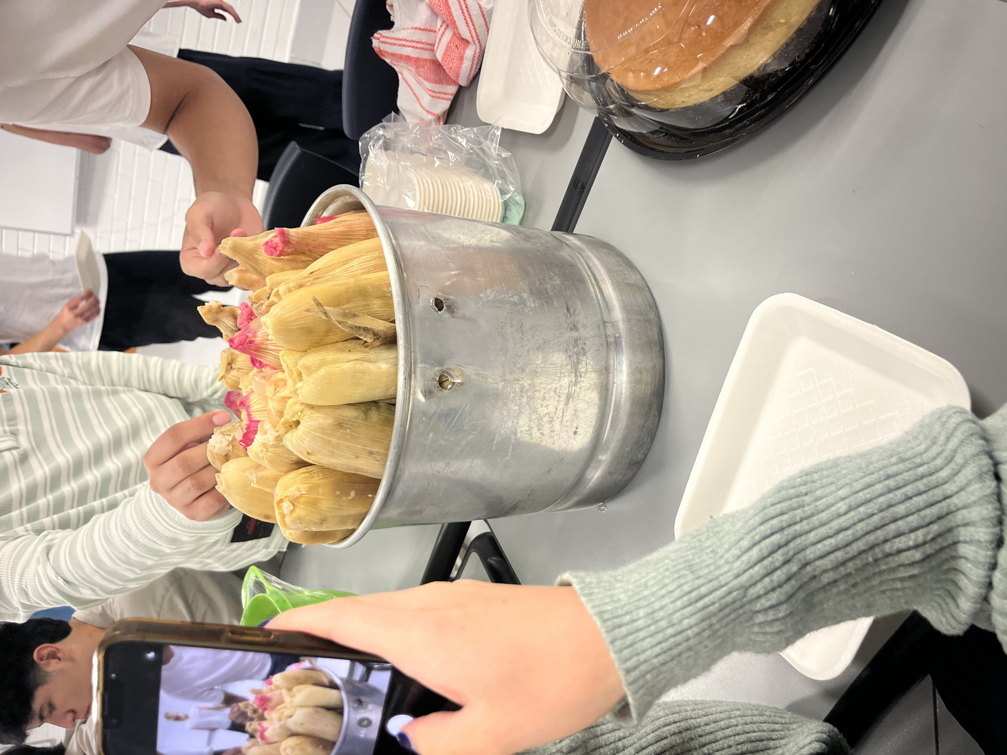 Here is a forensic description of the image:
The image captures an indoor scene, potentially a gathering or event in an unknown city. The focus is on a large, galvanized metal bucket filled to the brim with tamales, some revealing pink fillings. A person wearing a light green striped shirt is reaching into the bucket, presumably taking a tamale. A second person is using a smartphone to take a picture of the tamales, focusing on the bucket. A white plate is seen next to the bucket.
Additional elements on the table include a stack of white cups, a red and white striped cloth, and a cake under a plastic dome. Another person wearing a white shirt is partially visible. The environment appears to be well-lit, with a plain grey table as the surface.
The image documents an event where food, specifically tamales, is being served and photographed.