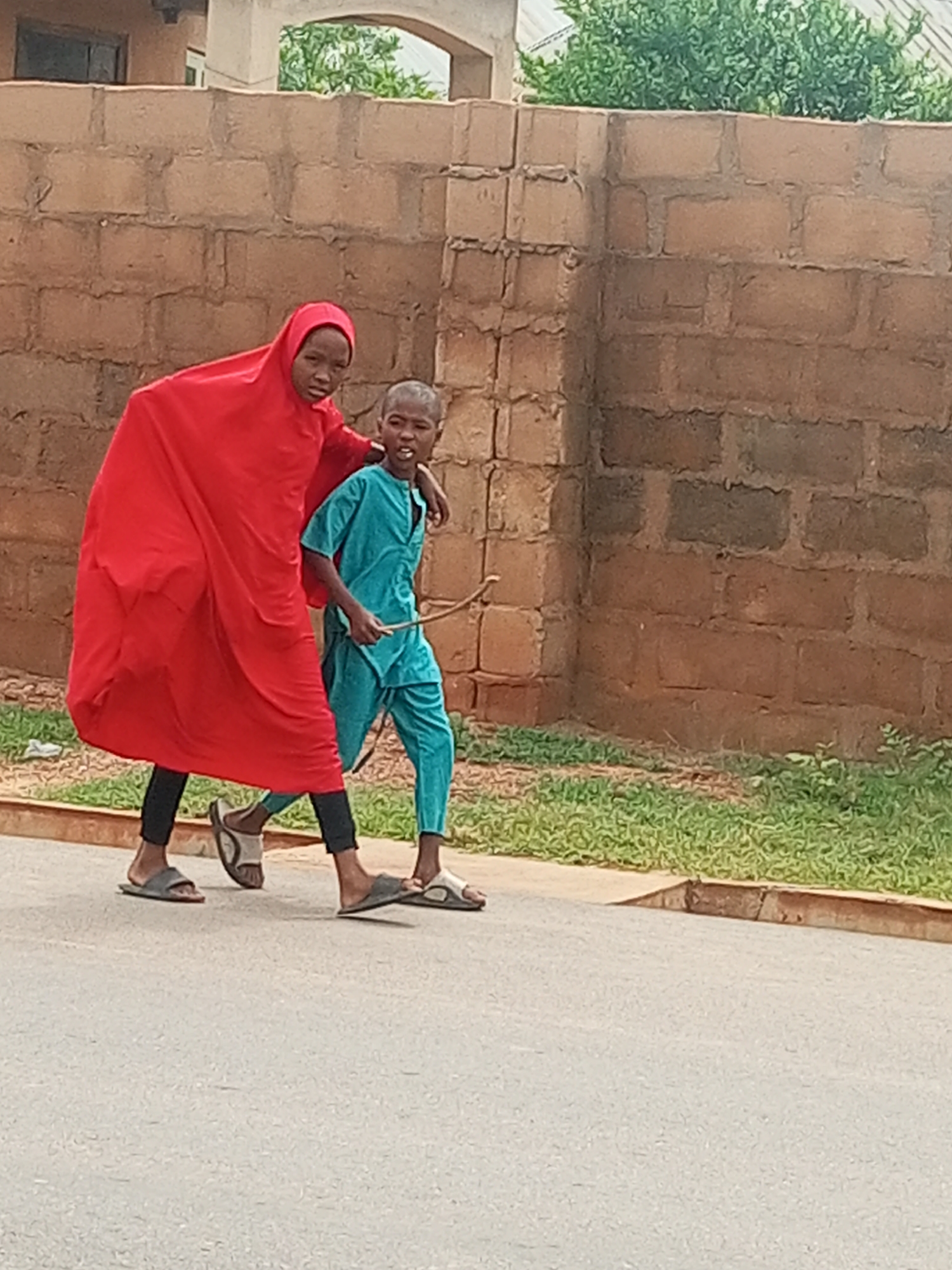 Two young people, one wearing a red hijab and a long dress, the other in a blue tracksuit, walk side-by-side on an asphalt road in front of a brown brick wall. The person in red appears to be guiding or supporting the person in blue, with their arm around their shoulder. The person in blue is holding a thin, curved object, possibly a stick or a small bow. Green grass and some weeds are visible along the edge of the road and behind the wall. The background includes a building with a window and a portion of a white structure with an arched opening. The scene appears to be outdoors in a residential or public area.