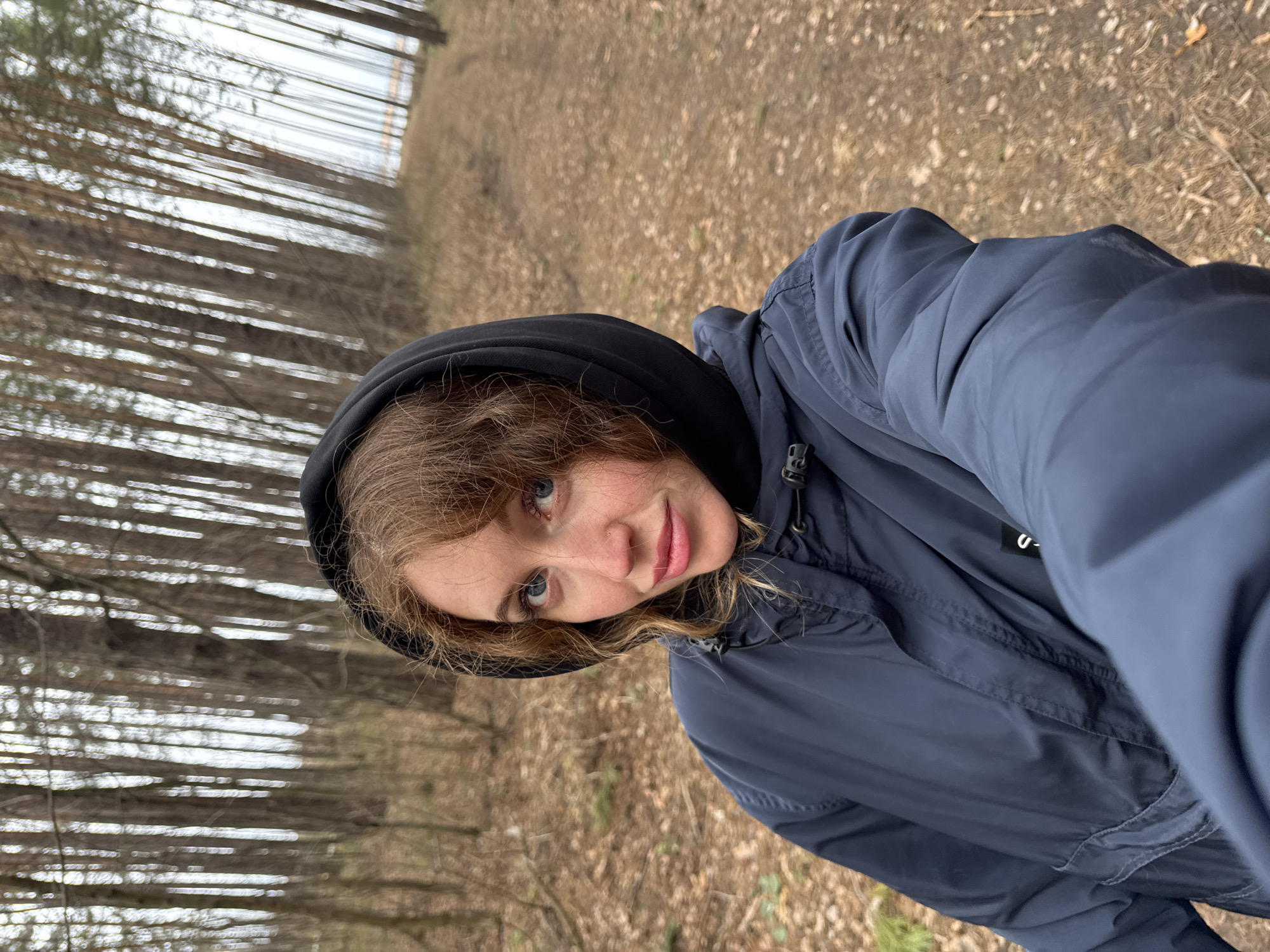 A young woman is taking a selfie in a forest. She is wearing a dark blue hooded jacket. The forest has tall, thin trees and the ground is covered with brown leaves and vegetation. The image suggests a natural environment, likely a wooded area in the city of Chaikovsky, Russia.