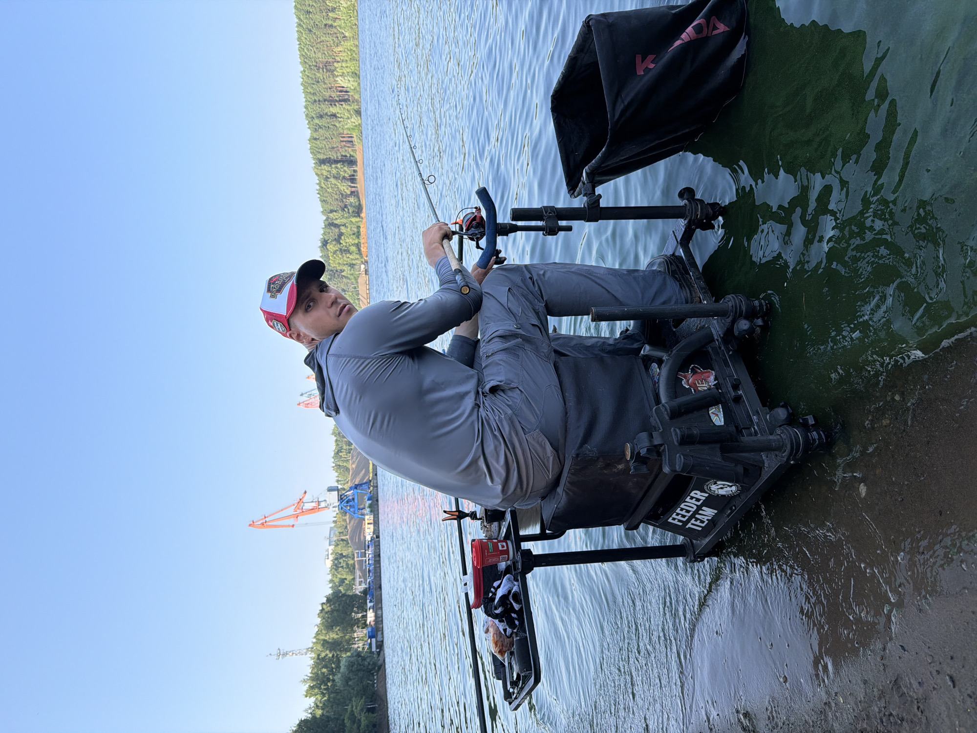 A young male is fishing from a feeder chair on the bank of the Kama River in Chaikovsky, Russia. He is wearing a grey long-sleeved shirt and grey fishing pants, along with a red and white baseball cap. The feeder chair is positioned at the water's edge, with a tray holding fishing accessories and a red cup. A black fishing net is attached to the chair. In the background, industrial cranes and a forest are visible on the opposite bank. The sky is clear and blue.