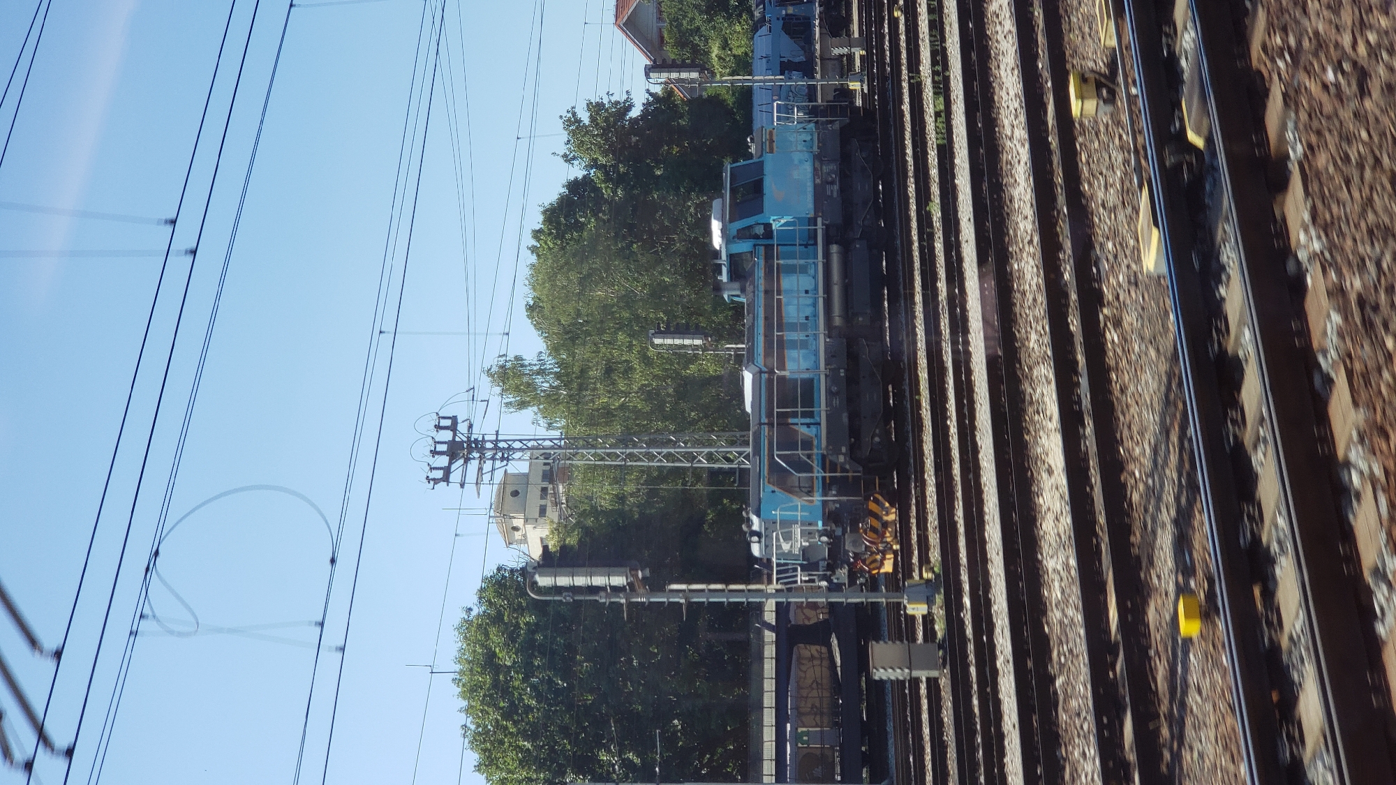 The image shows a train yard in Trnava, Slovakia. In the foreground, there are multiple parallel train tracks with ballast. A blue and gray diesel locomotive is visible in the middle ground, positioned on one of the tracks. Behind the locomotive, there are additional train cars and a large green tree line. In the background, there are buildings and overhead power lines characteristic of a railway infrastructure. The sky is clear blue, suggesting it is a daytime scene.