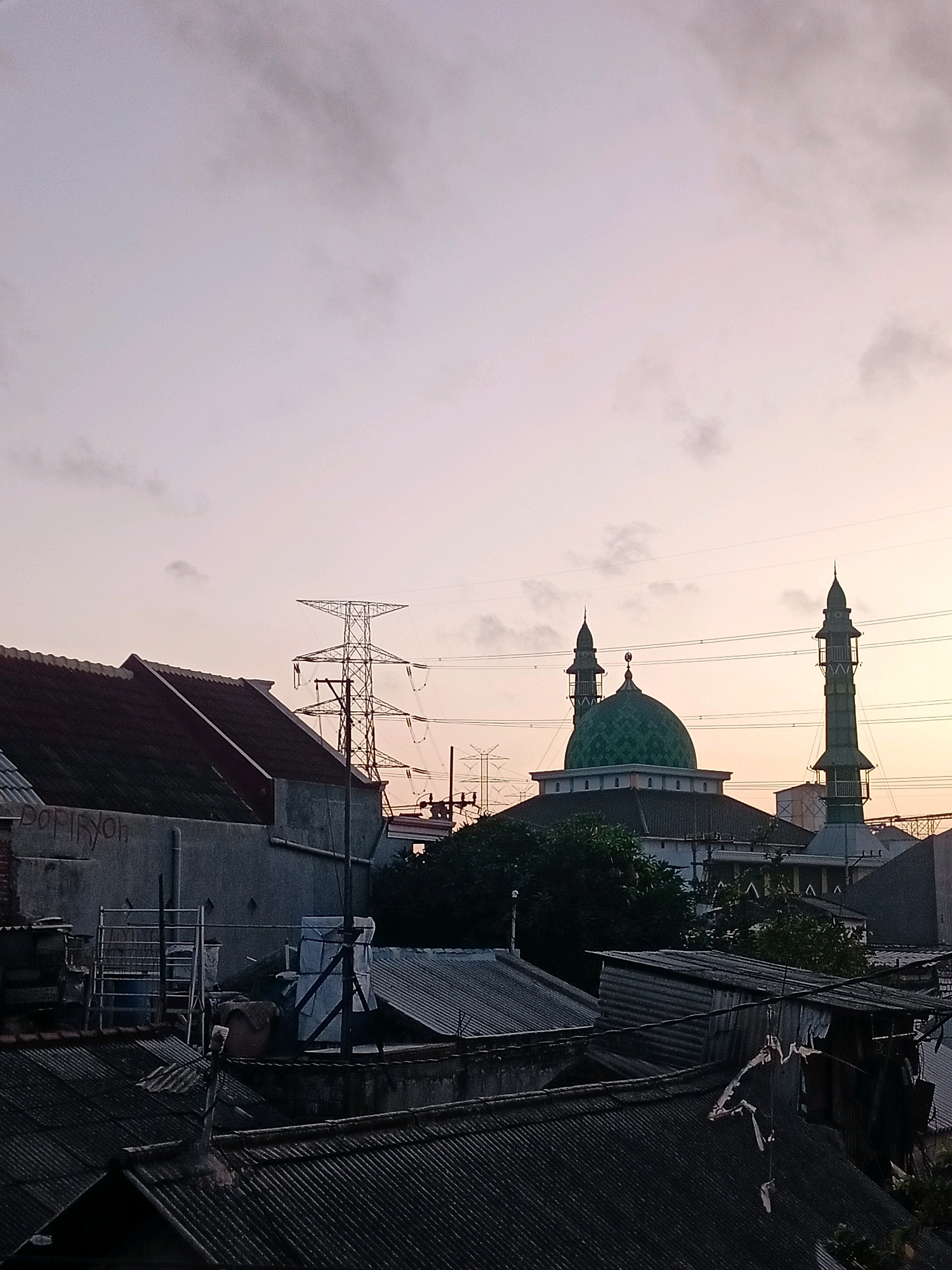 A photograph taken from a rooftop at dusk in Ngargosari, Indonesia, depicts an urban landscape dominated by residential rooftops and a prominent mosque. The sky, tinged with pale pink and lavender hues, indicates the setting sun. In the mid-ground, a green-domed mosque with two tall minarets stands out against the silhouette of other buildings and trees. To the left, a tall electricity pylon is visible, with power lines stretching across the frame. The foreground and mid-ground are filled with the corrugated metal roofs of various houses, some appearing weathered and in disrepair. The overall scene captures a quiet moment in a densely populated urban area as day transitions to night.
