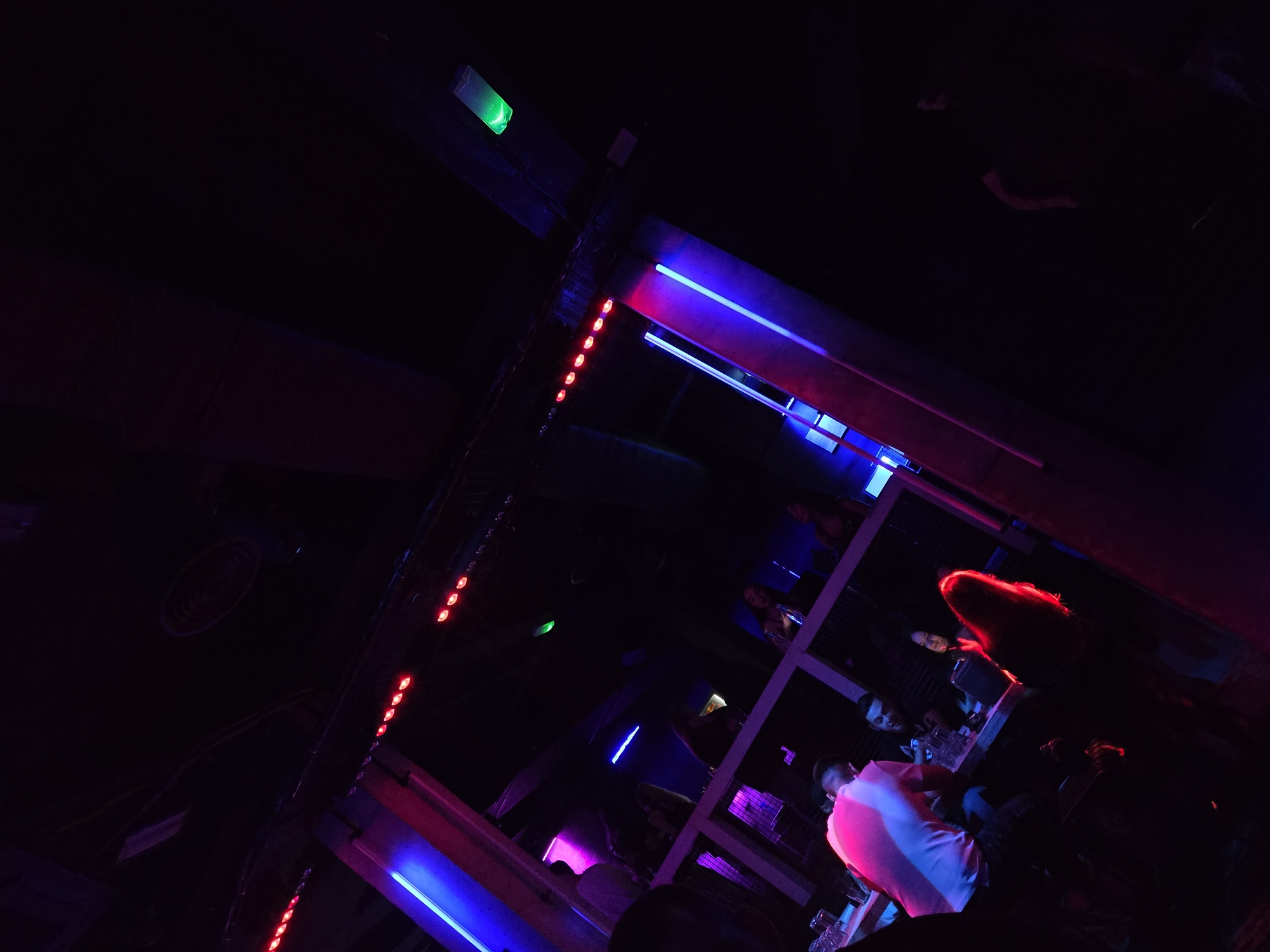 The image depicts the interior of a nightclub in Varna, Bulgaria. The scene is dimly lit with a combination of red, blue, and purple neon lights. A section of the nightclub's structural framework is visible in the foreground, adorned with a vertical row of red LED lights. A few people are seated at a table, with one wearing a light-colored shirt that reflects the pink lighting. A woman with bright red hair is also visible. The overall environment is consistent with a typical nightclub atmosphere.