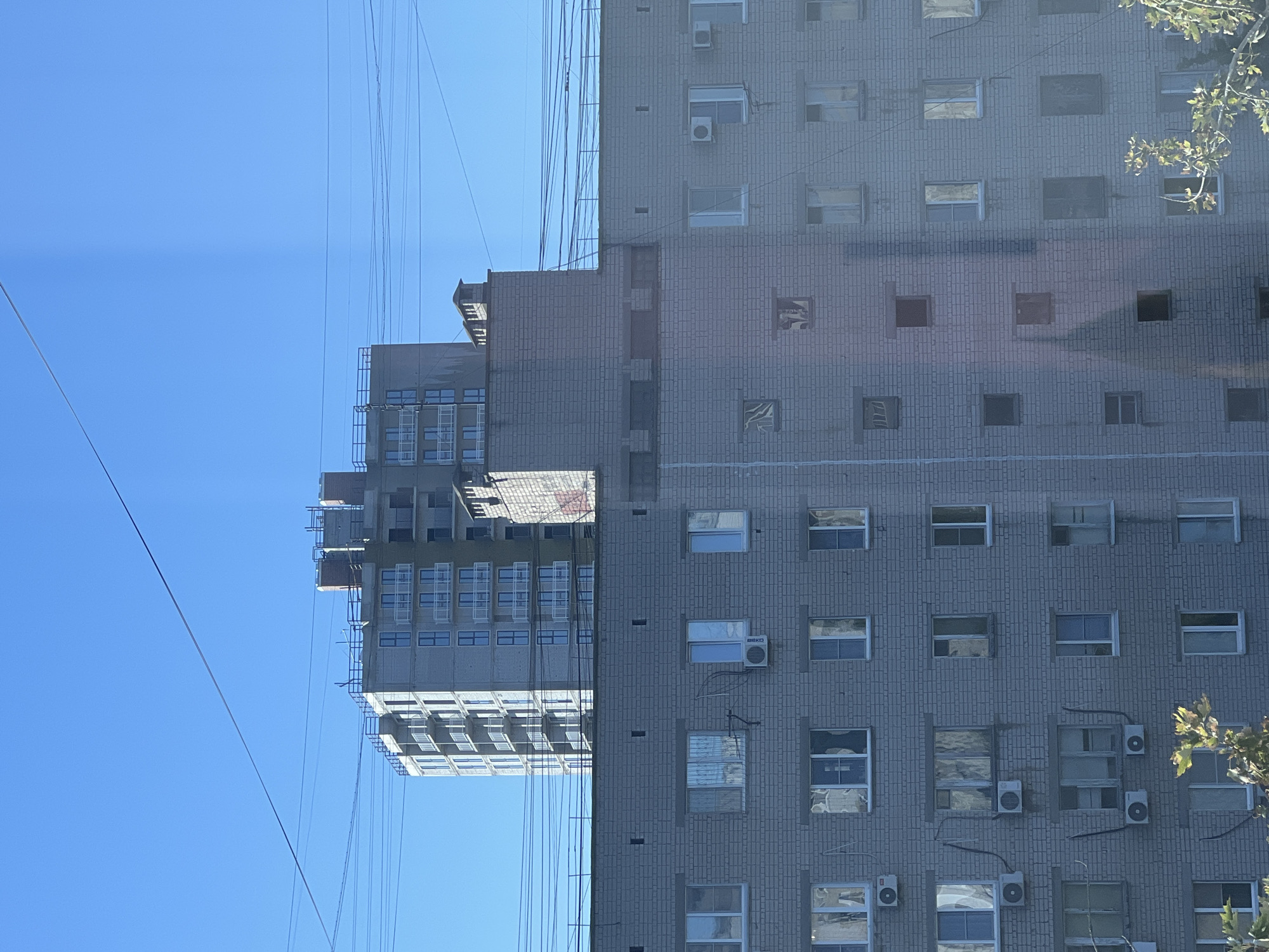 The image shows a view of a city skyline with several buildings under a clear blue sky. In the foreground, a multi-story building made of light gray brick is visible. It has numerous windows arranged in a grid pattern, with some windows appearing to have air conditioning units attached. Power lines crisscross the sky above this building. Behind the brick building, a taller, modern building under construction is visible. This newer building has a lighter facade and multiple balconies with white railings. The construction site at the top of this building indicates ongoing work. Another smaller brick structure, possibly part of a chimney or ventilation system, is situated between the two main buildings. The overall scene depicts an urban environment with a mix of older and newer construction, set against a bright, cloudless sky. This photograph was taken from an apartment window on the 6th floor of a residential building located at 98 Prospekt Lenina in Volgograd, Russia.
