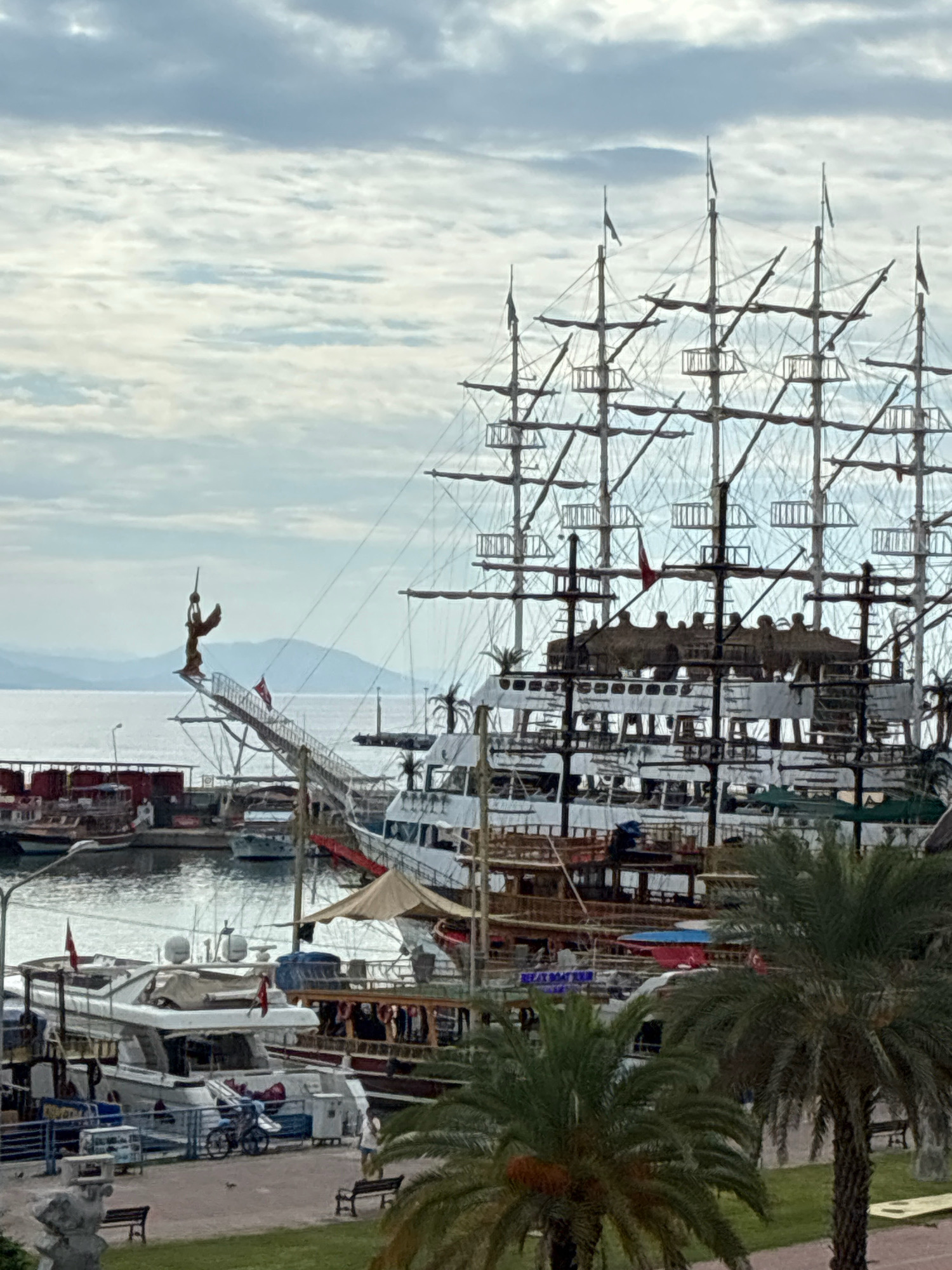 The image captures a harbor scene in Alanya, Turkey, dominated by a large, multi-masted sailing ship with numerous sails furled and rigging visible against a cloudy sky. The ship has several decks with thatched roofing and windows. A smaller, modern yacht is docked to the left of the large ship, with its sleek white hull and multiple antennas.
In the foreground, palm trees with lush fronds frame the scene. The waterfront area appears to be a promenade with benches and greenery. Other boats of various sizes are docked in the background, some appearing to be tour boats with multiple passengers visible on their decks. The water in the harbor is calm, reflecting the overcast sky. In the distance, hazy hills or mountains rise above the coastline, suggesting a coastal city location. A statue of a winged figure adorns the bow of the large sailing ship, positioned above a gangway leading to the shore.