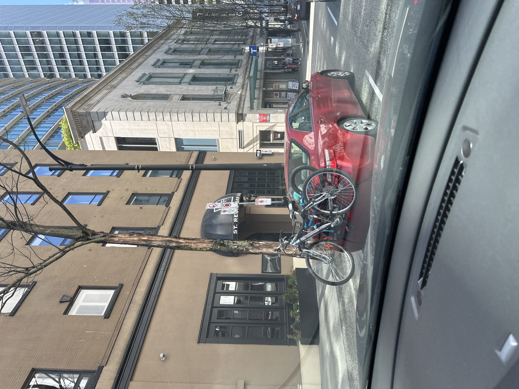 The image shows a street scene in Vancouver, Canada, viewed from inside a car. A red car is parked along the right side of the street, partially obstructing a bicycle rack in front of a building with a black awning marked "ST. REGIS BAR &." Another building is partially visible in the background, featuring a grid-like architectural pattern. A sidewalk is present in front of the buildings, and there are parking regulation signs posted near the bicycle rack. A tree branch extends across the facade of the building. The sky is blue and mostly clear.