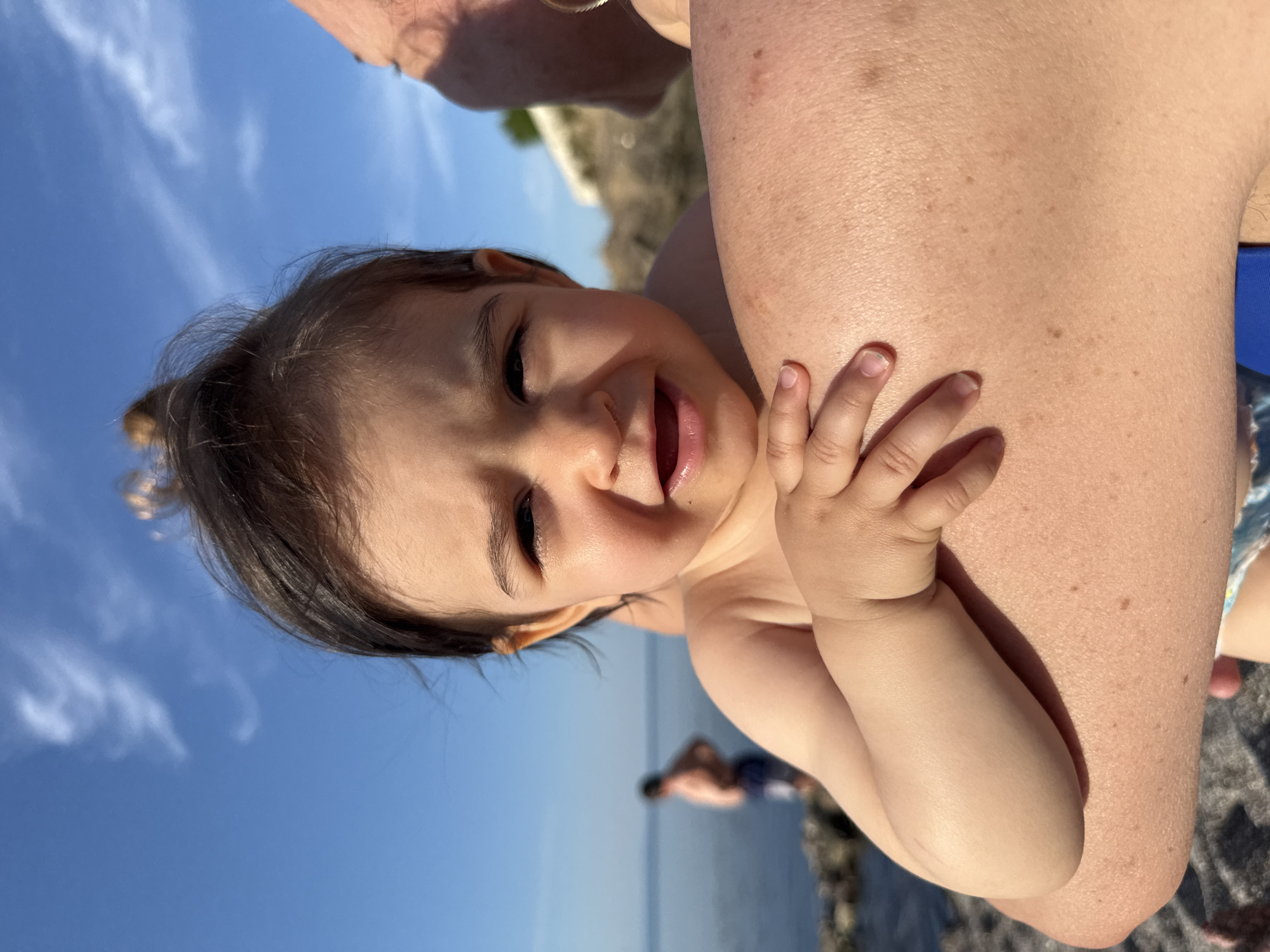 A close-up, eye-level, portrait-oriented shot focuses on the face of a baby, held by an adult whose arm is visible in the foreground. The baby, with dark hair styled in a bun, looks directly at the camera with wide eyes and an open mouth, appearing to be speaking or making a sound. The baby's left hand rests on the adult's arm. The background is blurred, showing a body of water, a rocky shoreline, and a person in the distance. The sky is clear blue with a few wispy clouds. The scene appears to be outdoors, likely at a beach or waterfront location, possibly in Sozopol, Bulgaria, given the context.