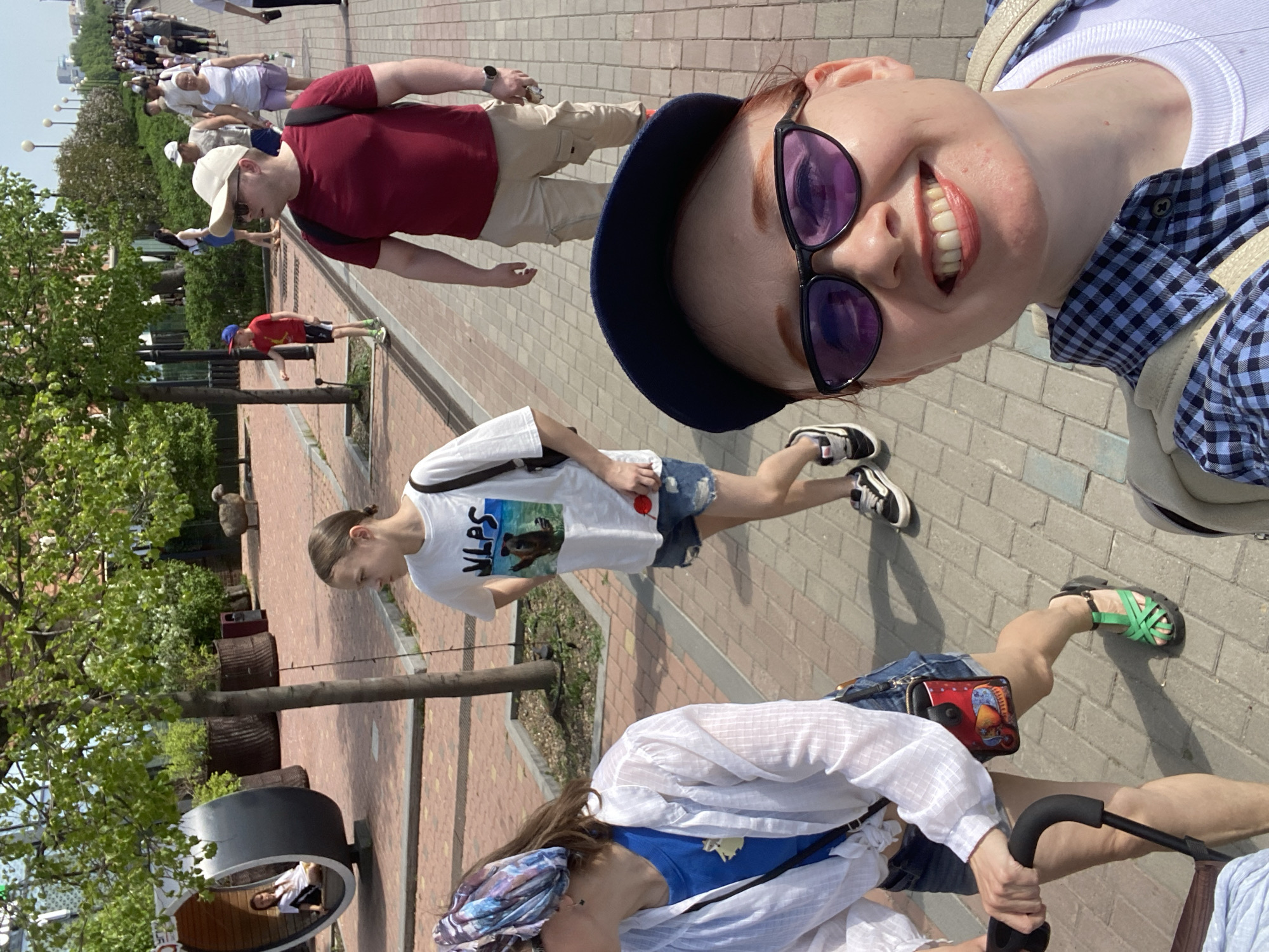 A medium shot shows a woman with auburn hair and fair skin, wearing a dark blue hat and dark purple sunglasses, smiling into the camera. She is positioned on the right side of the frame, with a checkered blue and white shirt partially visible. Behind her, a paved walkway with brick patterns stretches into the distance, occupied by numerous people walking in the same direction.
To the left, another woman walks, wearing a white t-shirt with a graphic on it, jean shorts, and sneakers. Further down the walkway, a man wearing a maroon t-shirt and tan pants is visible. In the lower part of the frame, another woman is pushing a stroller, wearing a white shirt, jean shorts, and colorful headwrap.
On the left side of the walkway, there are trees and cylindrical planters or containers, adding greenery to the scene. The environment appears to be an outdoor public space, potentially a park or promenade in a city setting. It is identifiable as the city of Yekaterinburg, Russia.
