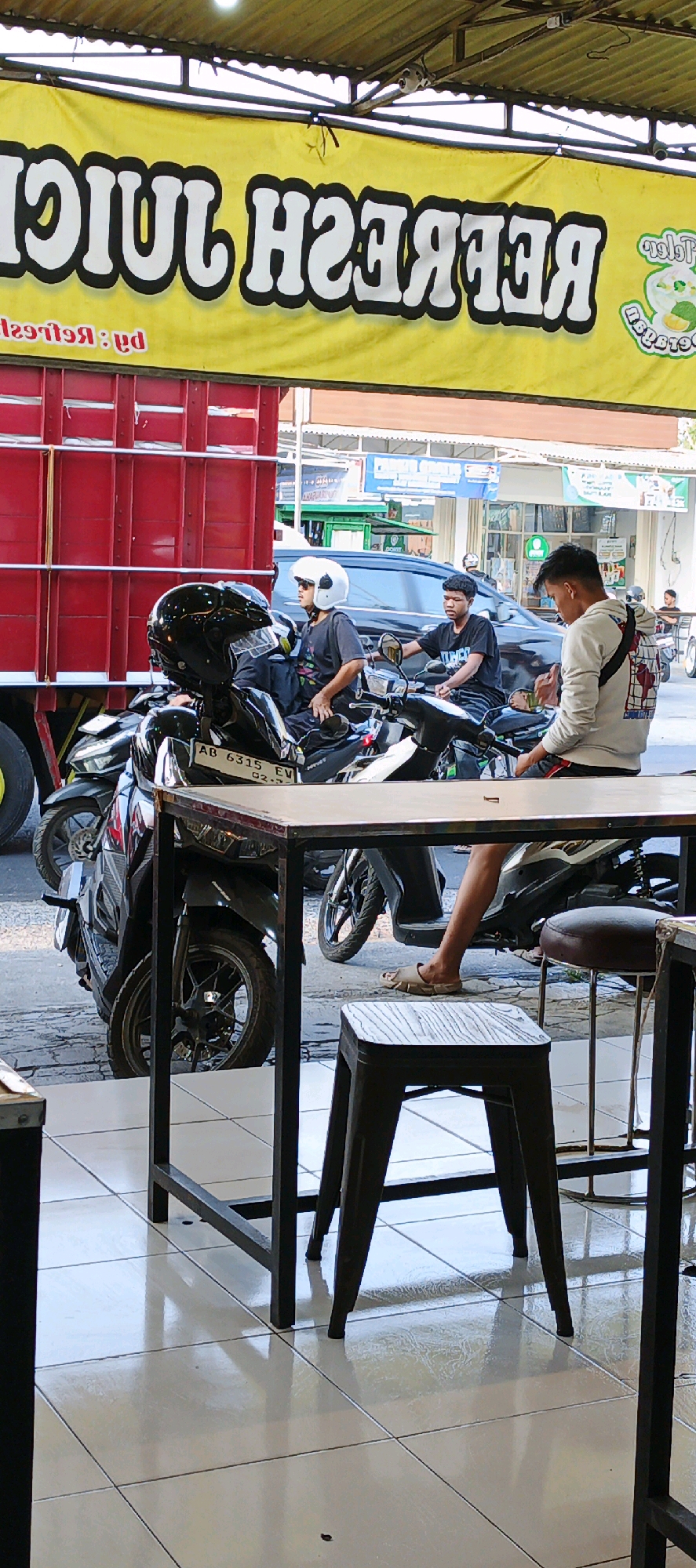 In Sidoarum, Indonesia, a street scene unfolds outside a juice vendor. A large red truck with a yellow banner advertising "REFRESH JUICE" is parked prominently. In front of the truck, several people are gathered around motorcycles. Two young men sit on separate motorcycles, one wearing a white helmet and sunglasses, the other looking forward. Another young man in a light-colored hoodie stands near the motorcycles, looking down. In the foreground, a metal table and a dark stool sit on a tiled floor, suggesting an outdoor seating area for the juice stand. The atmosphere appears casual and everyday, with vehicles and pedestrians typical of a bustling Indonesian street.