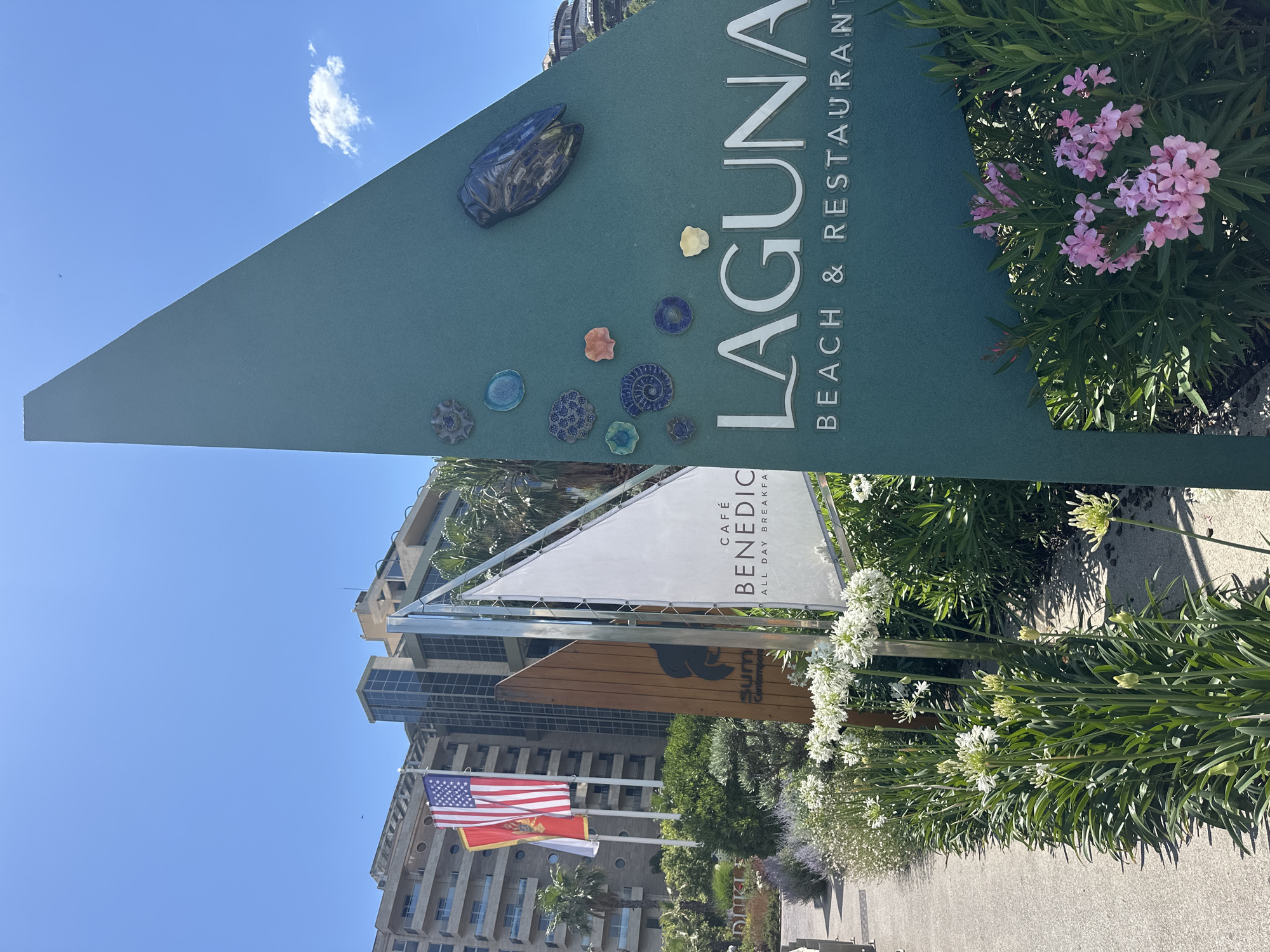 A large, teal green triangular sign stands prominently in the foreground, its surface decorated with several small, colorful ceramic or glass objects, including what appears to be a cicada figure. The sign displays white text that reads "LAGUNA BEACH & RESTAURANT". To the left of this sign, a white, sail-shaped sign bears the text "CAFÉ BENEDICT ALL DAY BREAKFAST". Further left, a wooden sign with a dark logo is partially visible. Behind these signs, American and Montenegrin flags fly from poles in front of a large, modern, multi-story building with balconies. The scene is bathed in sunlight, with a clear blue sky and a few scattered clouds. Lush green foliage and flowering plants, including white agapanthus and pink oleanders, surround the signs and the walkway, suggesting an outdoor setting. The overall impression is a sunny day at a seaside establishment in Budva, Montenegro.
