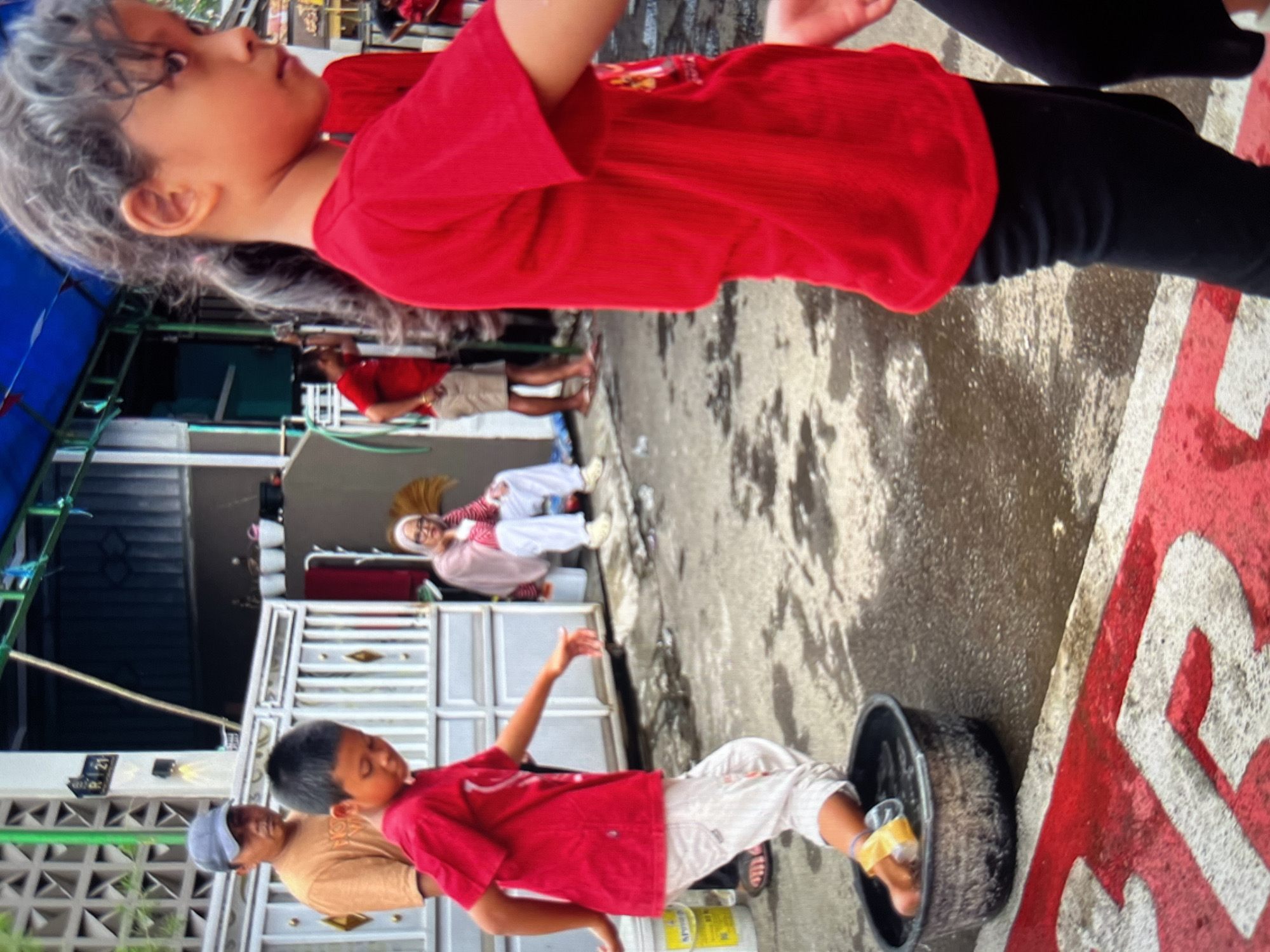 The image captures an outdoor scene in the city of Cirahayu, Indonesia, with a focus on children engaged in an activity. In the foreground, a young girl with dark hair pulled back in a ponytail, wearing a red t-shirt and black leggings, is positioned in profile, looking to the right. To her left, a boy in a red t-shirt and light-colored pants is stepping into a black basin filled with water. He appears to be balancing or playing a game. Behind him, an adult male wearing a tan t-shirt and a grey cap stands, observing the children. Further in the background, another adult male in a red t-shirt and tan shorts is visible. To the right of the first adult, an elderly woman wearing a pink headscarf and a light-colored outfit is seated. The environment appears to be a residential area, with a white fence and a building structure visible. The ground is wet and reflects the surrounding elements, suggesting recent rain or a water-related activity. Red painted markings are visible on the concrete ground in the foreground.
