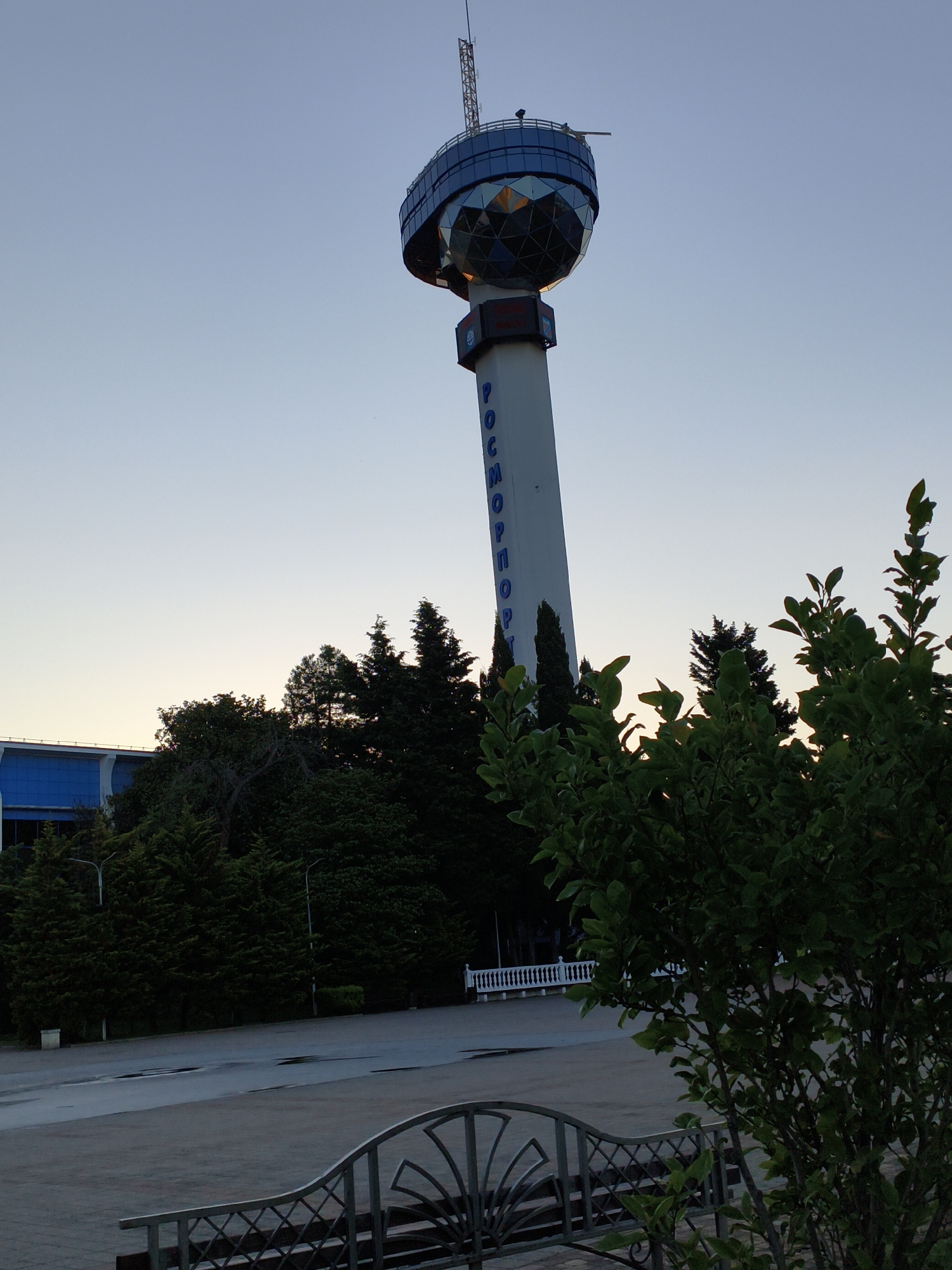 The photograph captures the "Rosport" tower in Agoy, Russia, against a clear, pale blue sky, likely at dawn or dusk. The tower, a prominent landmark, features a spherical observation deck at its apex, composed of many triangular glass panels that reflect the sky and surrounding lights. The tower's shaft is white and displays the blue Cyrillic lettering "РОСПОРТ" vertically.
In the foreground, to the right, a lush green shrub with broad leaves partially obscures the view. To the left of the tower, a modern building with a blue facade is visible, surrounded by a dense stand of dark green coniferous trees. Beyond the trees, a white ornate railing lines a walkway or plaza area, hinting at a public space. The ground in front of the trees appears to be paved, with a few puddles suggesting recent rain. A decorative metal fence with a curved design is also visible in the lower foreground. The overall impression is one of a quiet, possibly serene, outdoor scene at the edge of a populated area, dominated by the striking architecture of the tower.