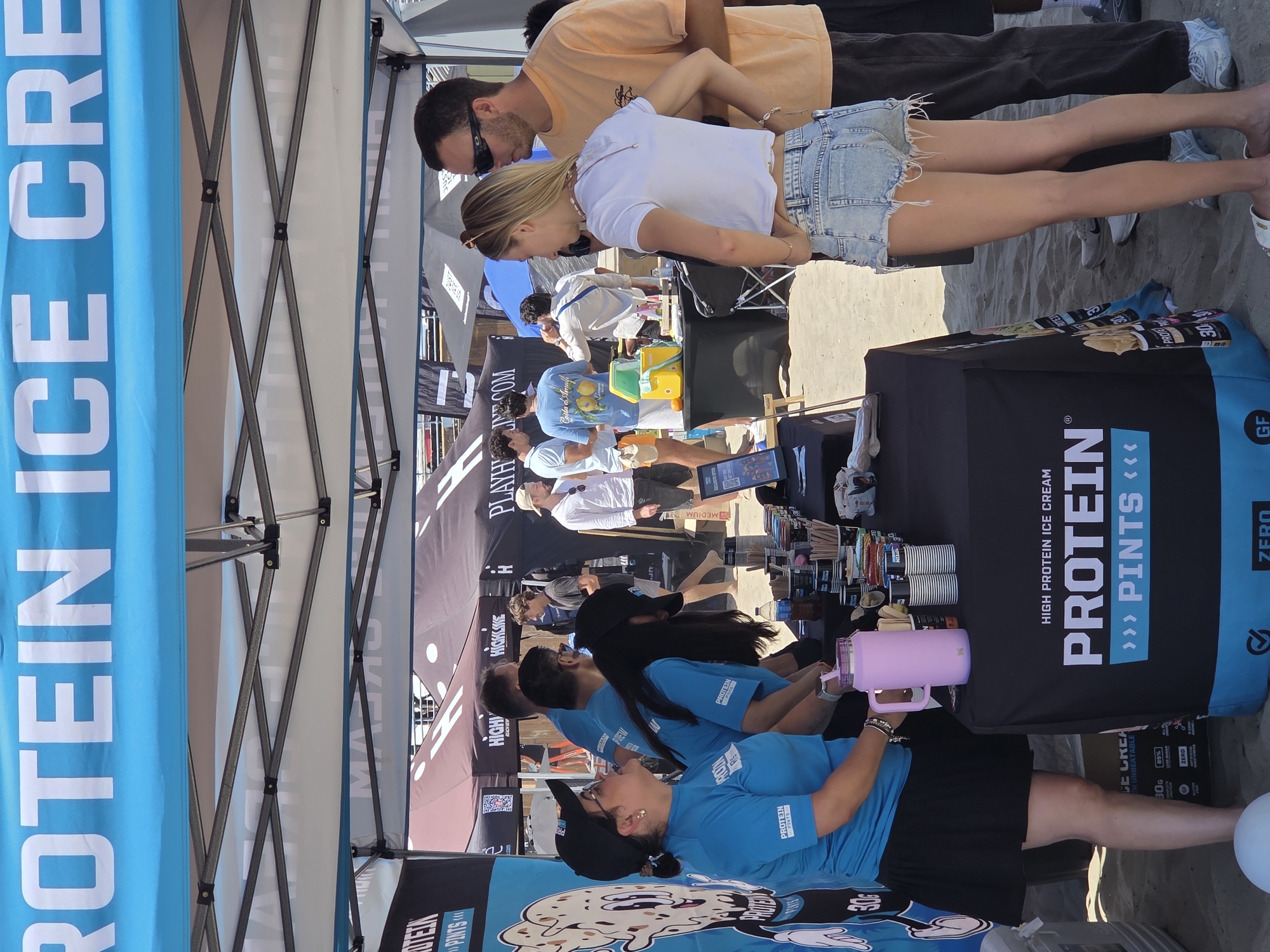 A group of people are gathered under a blue and white striped canopy on a sandy beach, likely at a promotional event for "Protein Ice Cream Pints." A woman wearing a blue shirt and black skirt is standing behind a table displaying various products and cups. To her right, a man and woman in casual beach attire are conversing. In the background, other individuals and tents are visible, suggesting a lively outdoor event. The overall scene conveys a relaxed, summery atmosphere.