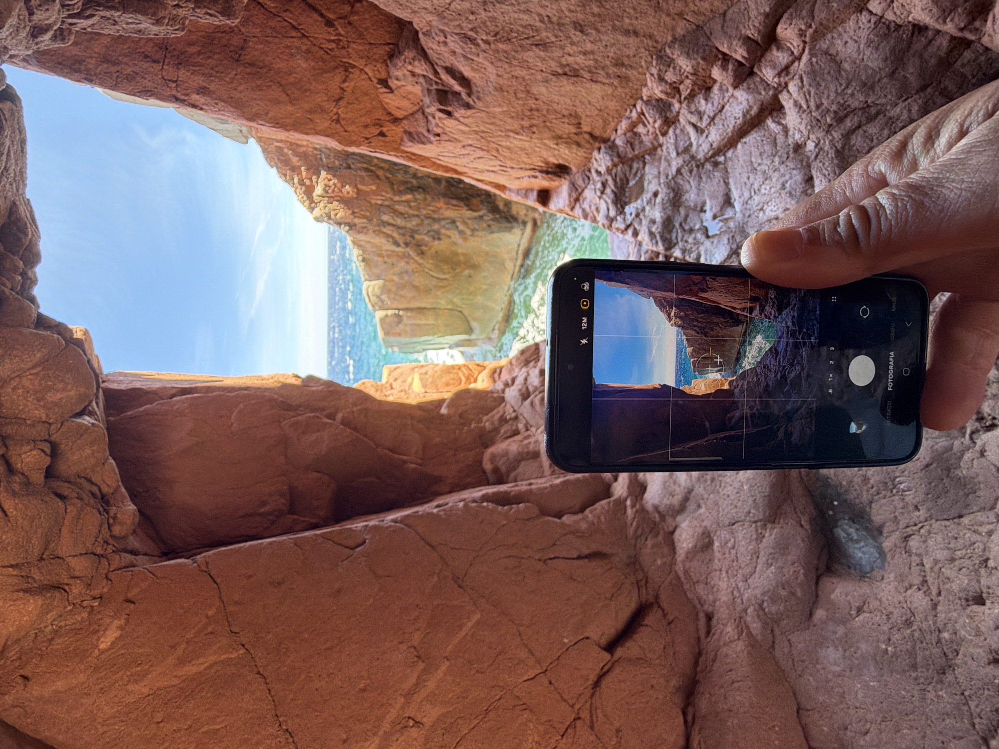 A hand holds a smartphone, capturing a view through a rock archway. The arch frames a seascape with the bright blue sky above and the turquoise ocean below. The rock formations are reddish-brown and textured, with visible cracks and striations. The phone screen displays a photo of the same scene, mirroring the view through the arch. The overall environment suggests a coastal cave or grotto, likely in a Mediterranean location such as Tortolì, Italy, known for its rocky coastline and coves.