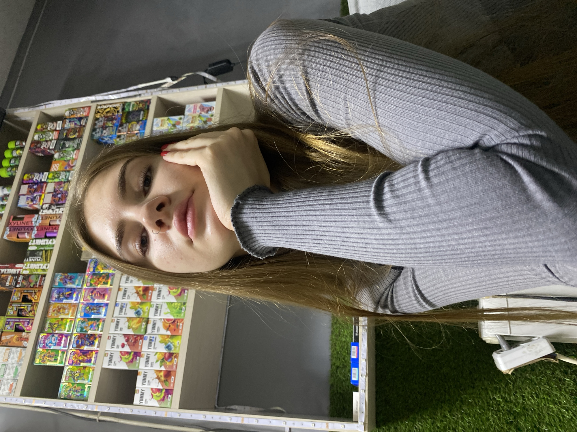 A young woman with long, light brown hair is pictured in a dimly lit room. She is looking towards the camera with her right hand propping up her chin and her left arm resting on her chest. She is wearing a grey ribbed sweater. Behind her, shelves are stacked with various colorful boxes, possibly e-liquid or vape products, and some green artificial grass is visible on the lower shelves. The scene appears to be indoors, within a commercial establishment such as a vape shop. The location, Дьяково, Russia, does not provide any additional visual context to the image itself.