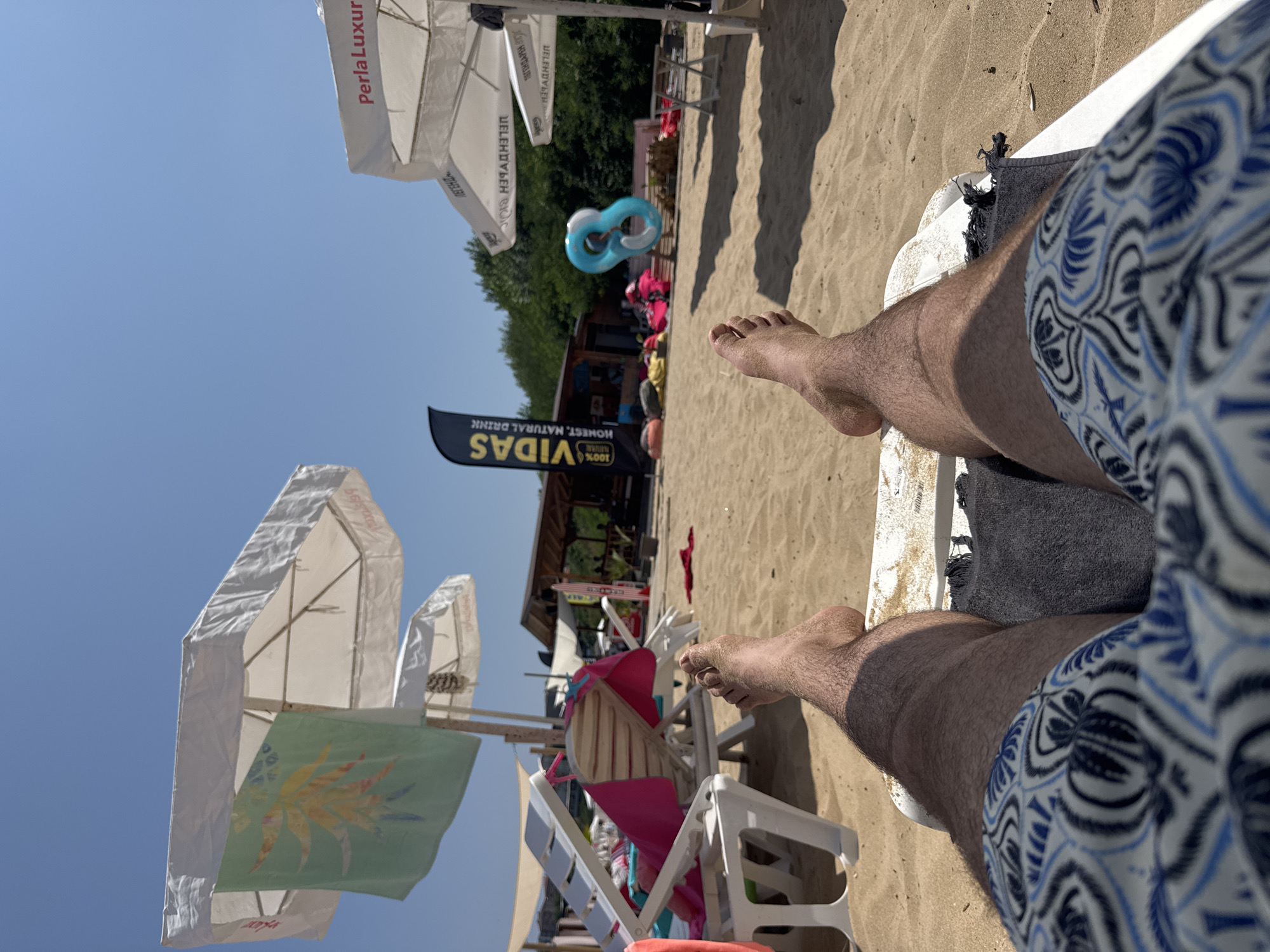 The image captures a first-person perspective of a person relaxing on a beach chair on a sandy beach. The person is wearing patterned swim trunks and their feet are visible, covered in sand. In the background, there are several beach umbrellas, some of which are white and appear to have writing on them. A green towel with a yellow and orange leaf design is hanging from one of the umbrellas. There are also other beach chairs, some with pink towels. Further back, a wooden structure with a roof, possibly a beach bar or cafe, is visible, with trees and foliage behind it. A tall black banner with yellow text reads "VIDAS 100% HONEST. NATURAL DRINK." A blue inflatable ring is floating in the air, seemingly thrown. The sky is clear and blue. The scene is set on a sunny day at a beach resort in Primorsko, Bulgaria.