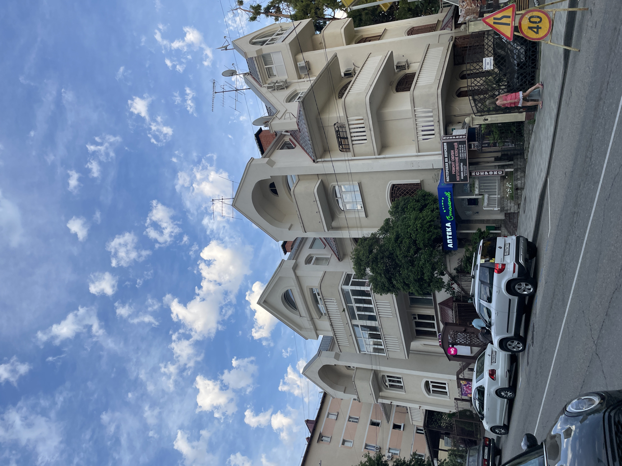 The image shows a street scene in Sochi, Russia, with a modern, multi-story building on the right and a lighter colored apartment building on the left. The sky is blue with scattered white clouds. In front of the building on the right, there are several cars parked along the street, including a white SUV and a white sedan. A woman in a pink and white dress is walking on the sidewalk. There are also several signs visible, including one with a speed limit of 40. The building on the right has balconies, arched windows, and several antennas on the roof. A tree with dense green foliage is growing next to the building. The building on the left appears to be an older style apartment building with multiple windows.