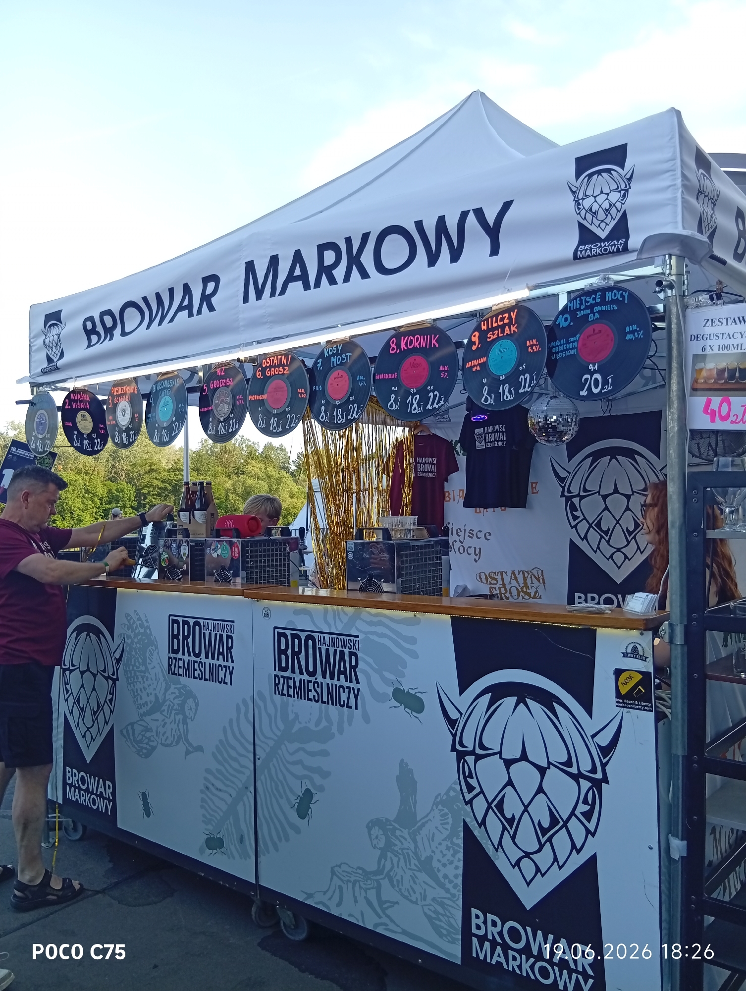 The image shows an outdoor beer stand with a white tent canopy that has the text "BROWAR MARKOWY" printed on it. Several circular signs resembling vinyl records, each with beer names and prices, are hanging from the canopy. A man in a maroon t-shirt is standing behind the counter, and a young person with blonde hair is partially visible behind him. The counter displays bottles of beer, a JBL speaker, and other items. The side of the stand is decorated with graphics of hops and other designs, along with the text "HAJNOWSKI BROWAR RZEMIEŚLNICZY" and "BROWAR MARKOWY". A disco ball hangs from the canopy. The background shows trees and foliage, suggesting an outdoor setting, possibly a festival or event. The bottom left corner of the image displays "POCO C75" and the bottom right shows "19.06.2026 18:26". It is not possible to determine the exact location in Wrocław, Poland from the image.