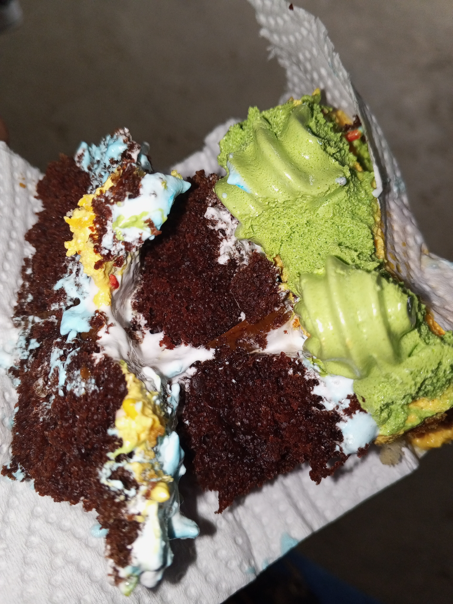 The image shows a piece of chocolate cake held in a white paper towel. The cake is decorated with green, yellow, blue, and white frosting. The location and other details are unknown.