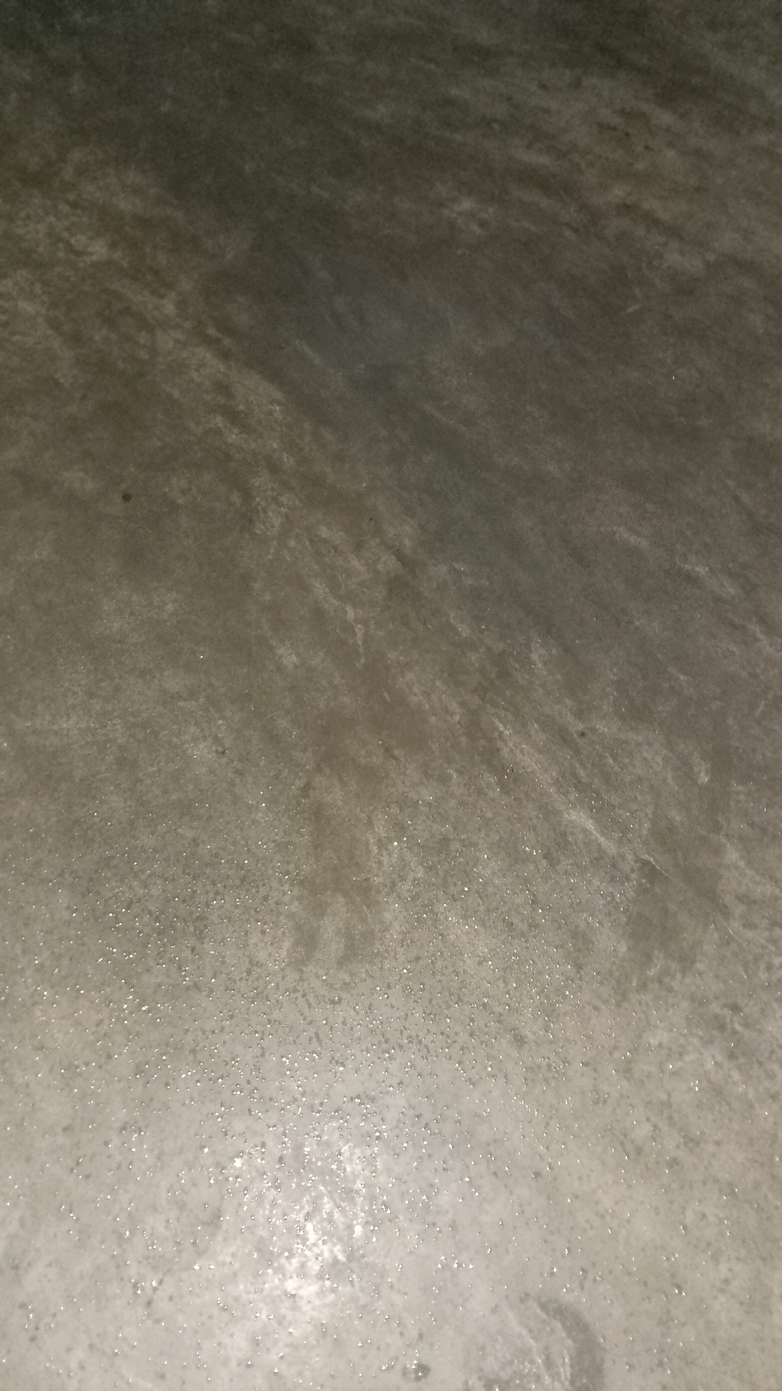 The image shows a close-up, oblique view of a textured, wet concrete surface. The surface has a gradient from darker, drier concrete at the top to lighter, wet concrete with visible water droplets and reflections at the bottom. There are no discernible people, subjects, or specific objects in the frame. The environment appears to be an outdoor or semi-outdoor concrete floor. The location in Mulyo Haji, Indonesia, is not visually evident from the image itself.