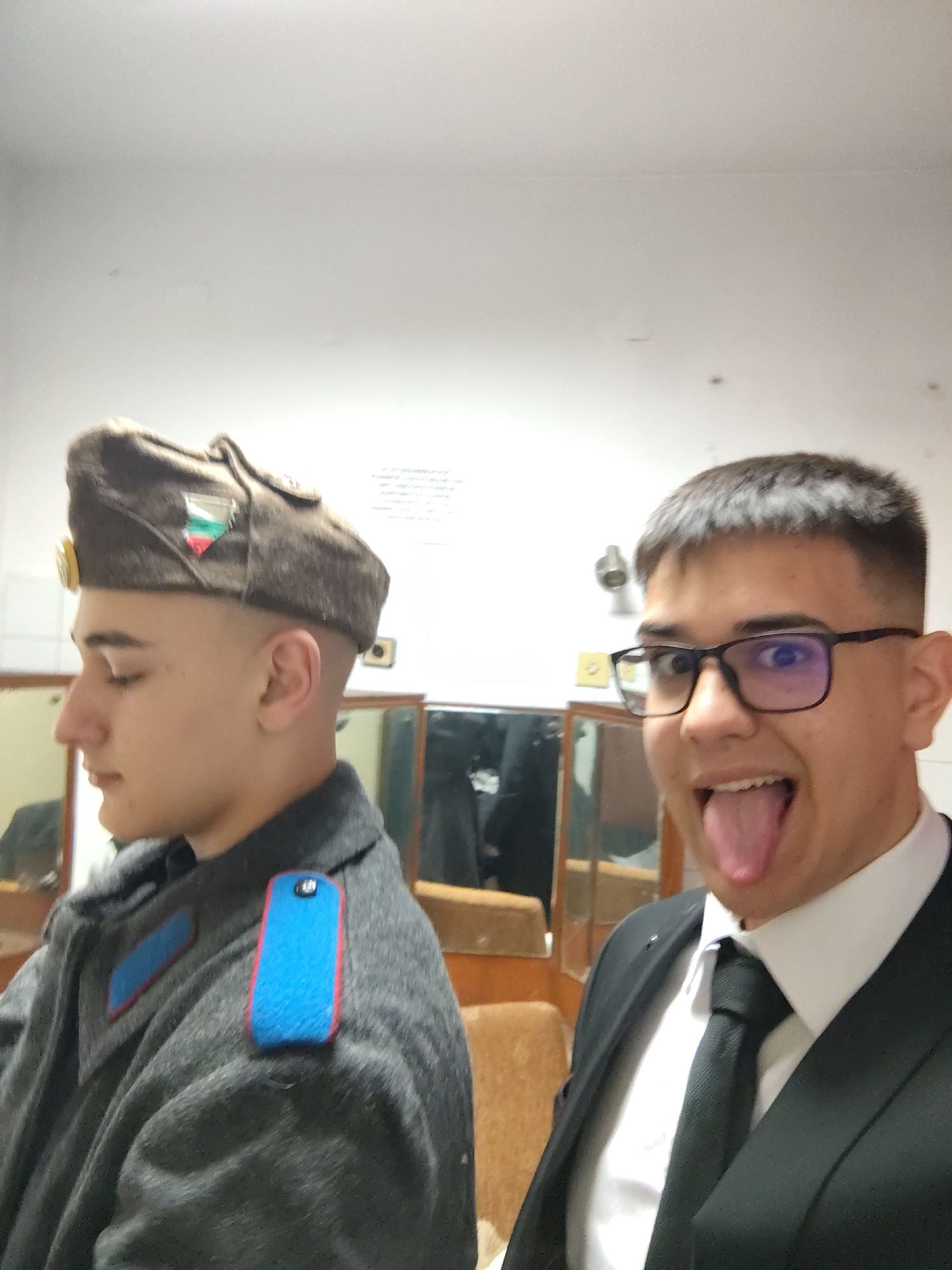Two young men are indoors. The man on the left wears a military-style uniform with a cap bearing the Bulgarian flag. The man on the right wears a suit and tie, and glasses, and is sticking out his tongue. The setting appears to be a room with mirrors. The city and specific location cannot be determined from the image.