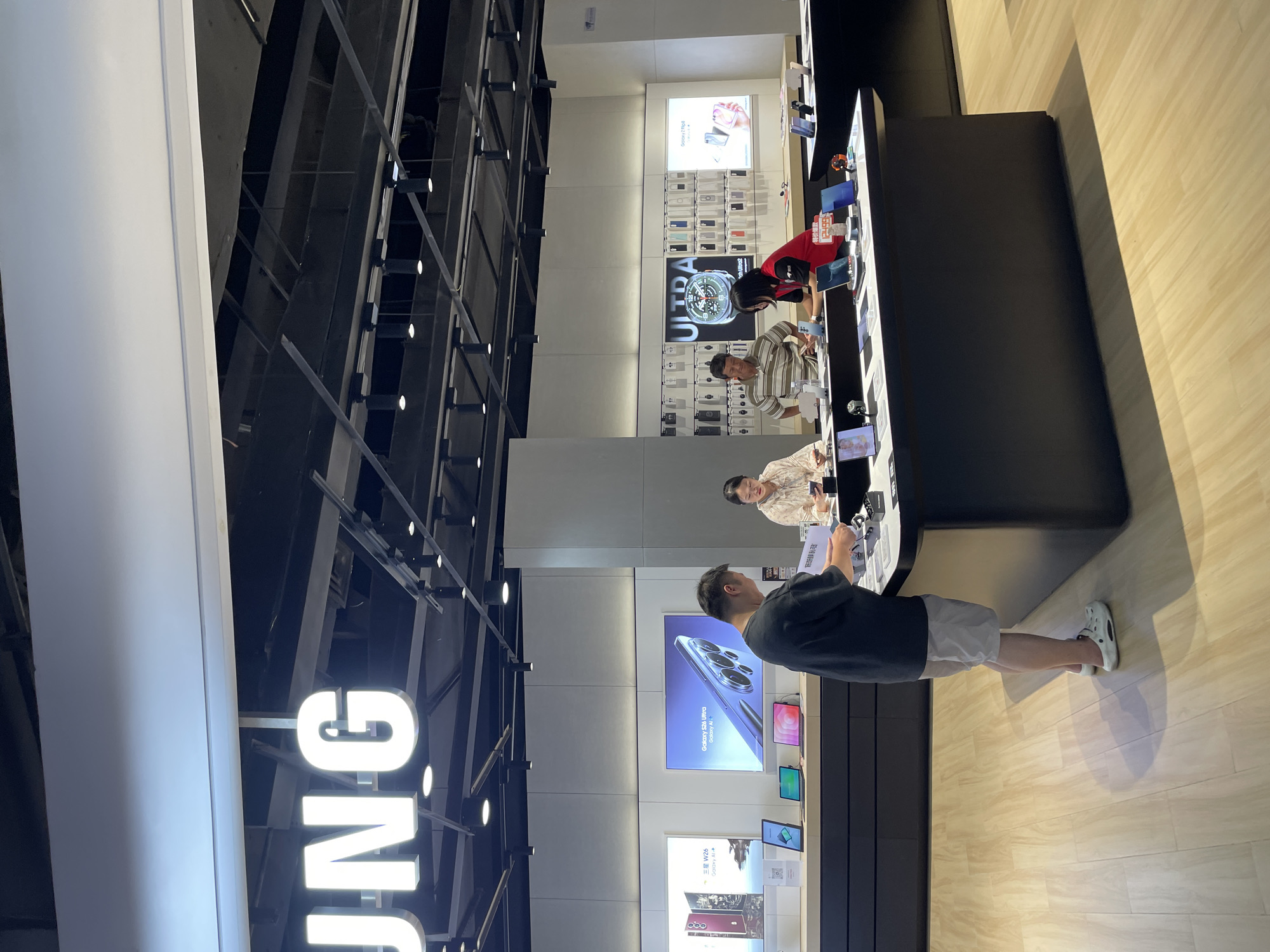 In a Samsung store in Liangjiang, China, a male customer in a dark t-shirt and light-colored shorts, wearing white Crocs, stands at a black counter, examining some paperwork. Behind him, on display shelves, are various Samsung phone cases and devices. To his left, a large screen displays a Samsung phone advertisement. To the right of the counter, a female employee in a red vest assists a male customer in a striped shirt. Another female employee in a floral shirt is also present. The store features a light-colored wooden floor and modern lighting fixtures.