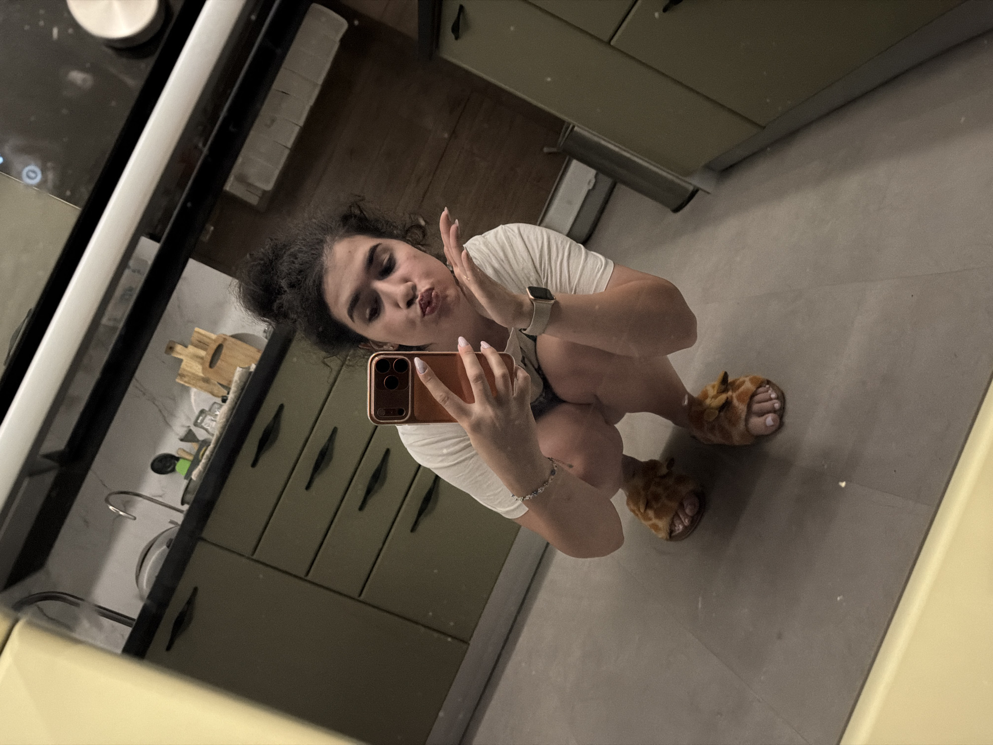 A young woman is photographed in a kitchen, possibly in Sofia, Bulgaria. She is squatting down and taking a selfie with her phone. She is wearing a white t-shirt, denim shorts, and furry animal-themed slippers. Her hair is pulled up in a messy bun, and she is puckering her lips. A silver bracelet is visible on her left wrist, and a smartwatch is on her right wrist. The kitchen appears modern, with olive-green cabinets and stainless steel appliances. The floor is tiled in a neutral gray. The lighting suggests it is nighttime or indoors with artificial light.
