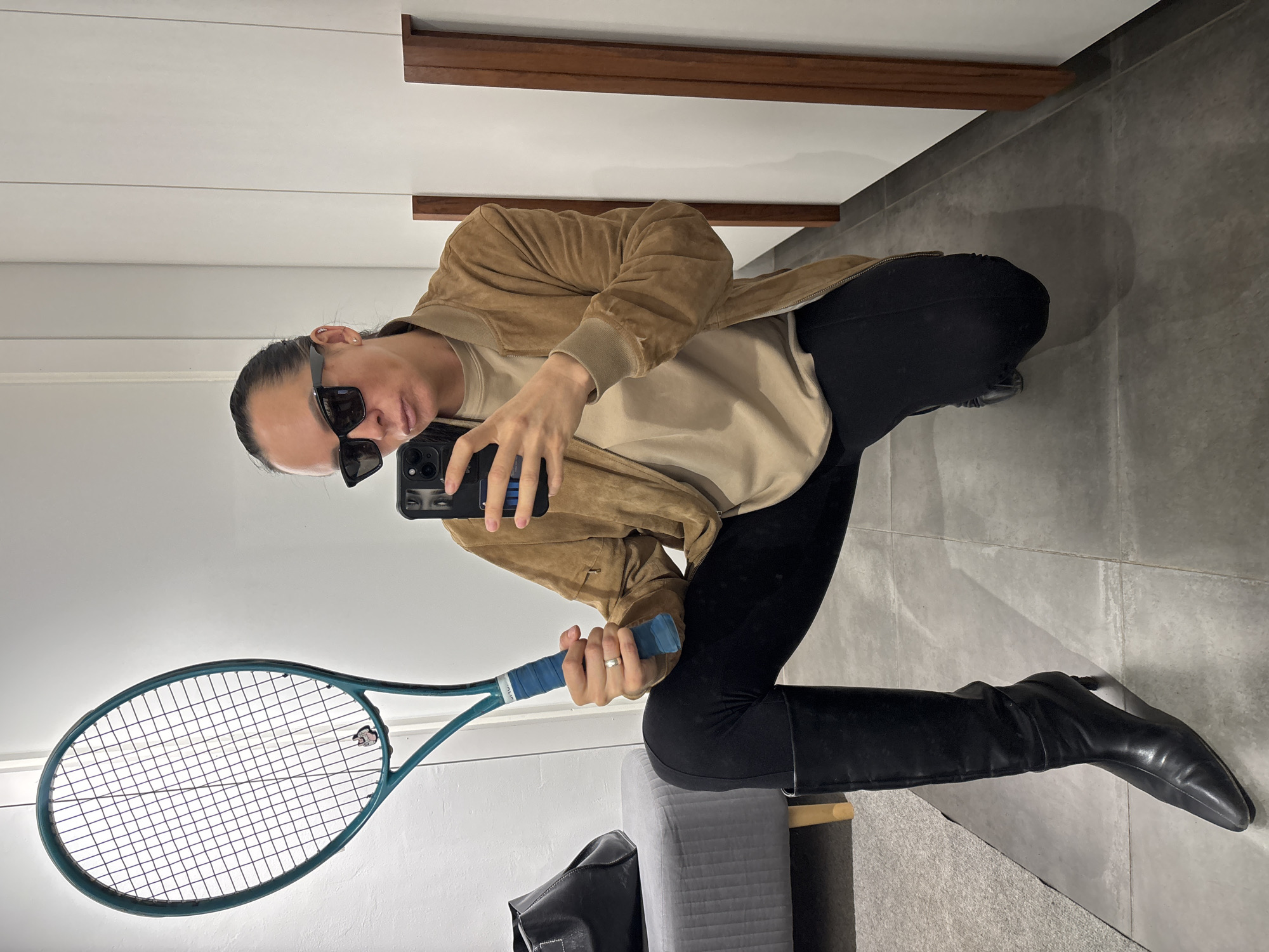 A woman is crouching in front of a mirror, holding a tennis racket and a smartphone. She is wearing black leggings tucked into black knee-high boots, a beige sweatshirt, and a tan suede jacket. She has sunglasses on and appears to be taking a selfie. The background includes a tiled floor, a wooden trim, a white wall, a gray stool, and a black bag. The location cannot be determined to be Chaikovsky, Russia from the image.