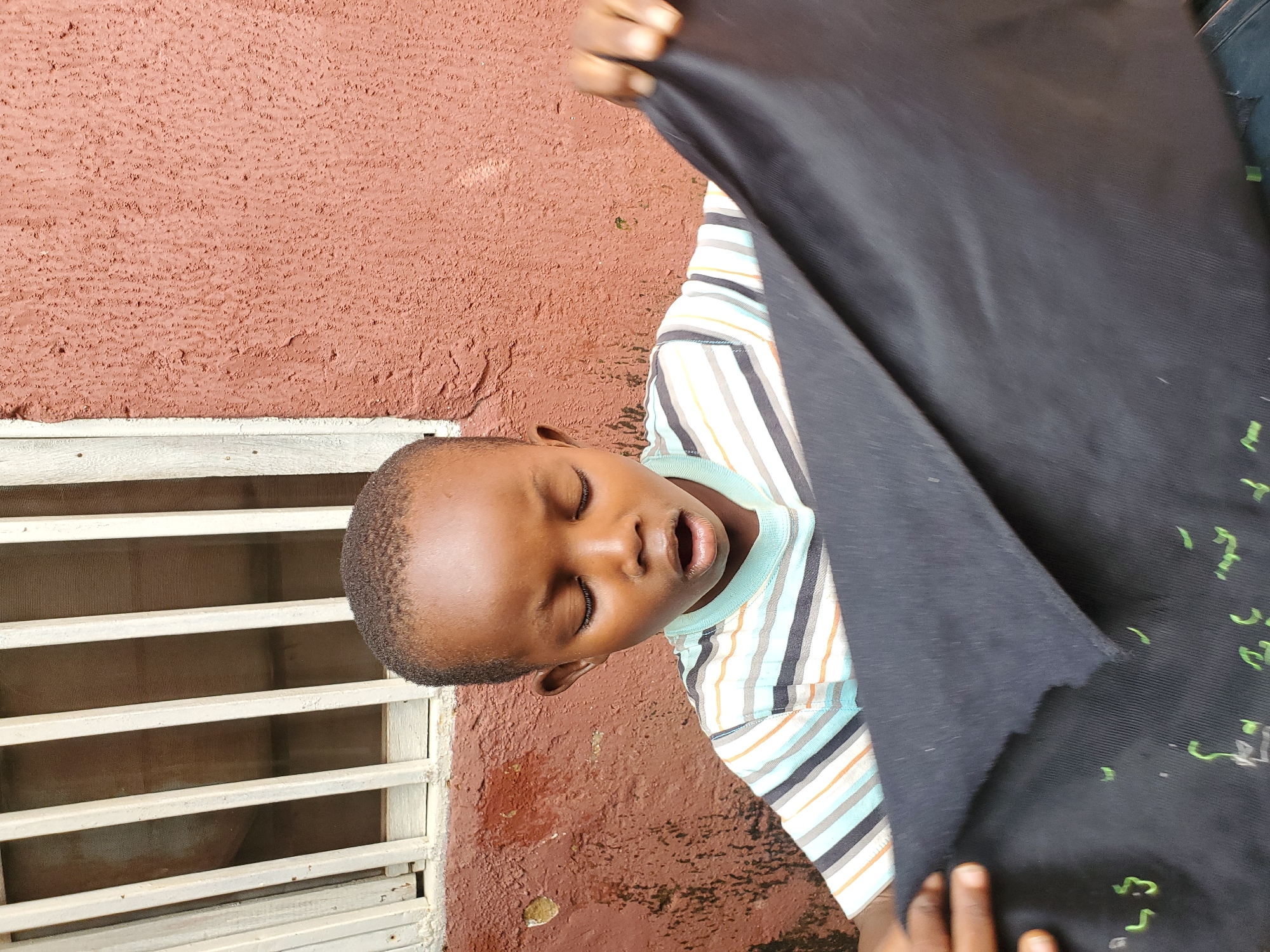 The image shows a young boy, likely of African descent, leaning against a rough, reddish-brown textured wall. He is wearing a striped t-shirt with colors including white, blue, orange, and black. The boy's eyes are closed, and his mouth is slightly open, suggesting a moment of rest or perhaps a playful pose. His head is tilted back.
In front of the boy, a large piece of dark, possibly black, fabric is held up, obscuring part of his body and the background. The fabric has some faint green markings on it, though they are difficult to decipher. The boy's hands are visible, holding the fabric.
To the left of the boy, a section of a window or ventilation grille is visible. It consists of horizontal white slats with a fine mesh behind them. The wall itself appears weathered, with some darker patches and a general texture that suggests an outdoor setting.
The overall lighting suggests it is daytime, possibly late morning or early afternoon, given the brightness and the shadows. The combination of the wall texture, the window grille, and the clothing hints at a casual, possibly domestic, outdoor environment in a warm climate. There is no visible text in the image.