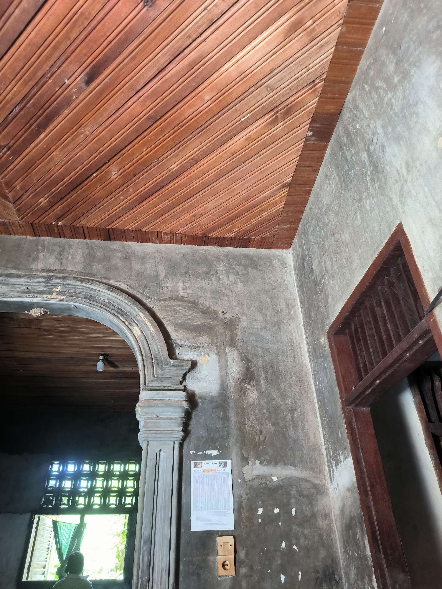 The image presents an interior view of a building in Merbopuntong, Indonesia, characterized by aging infrastructure and a mix of architectural styles. The ceiling is covered in wooden planks arranged diagonally. The walls are a faded, weathered gray with signs of deterioration and patching. An arched doorway with decorative molding stands on the left side of the frame. A calendar and electrical outlets are mounted on the wall near the doorway. A window with ornate latticework is visible through the doorway. The bottom left shows the back of a person looking out the window. A dark-framed window is positioned on the right side of the image.