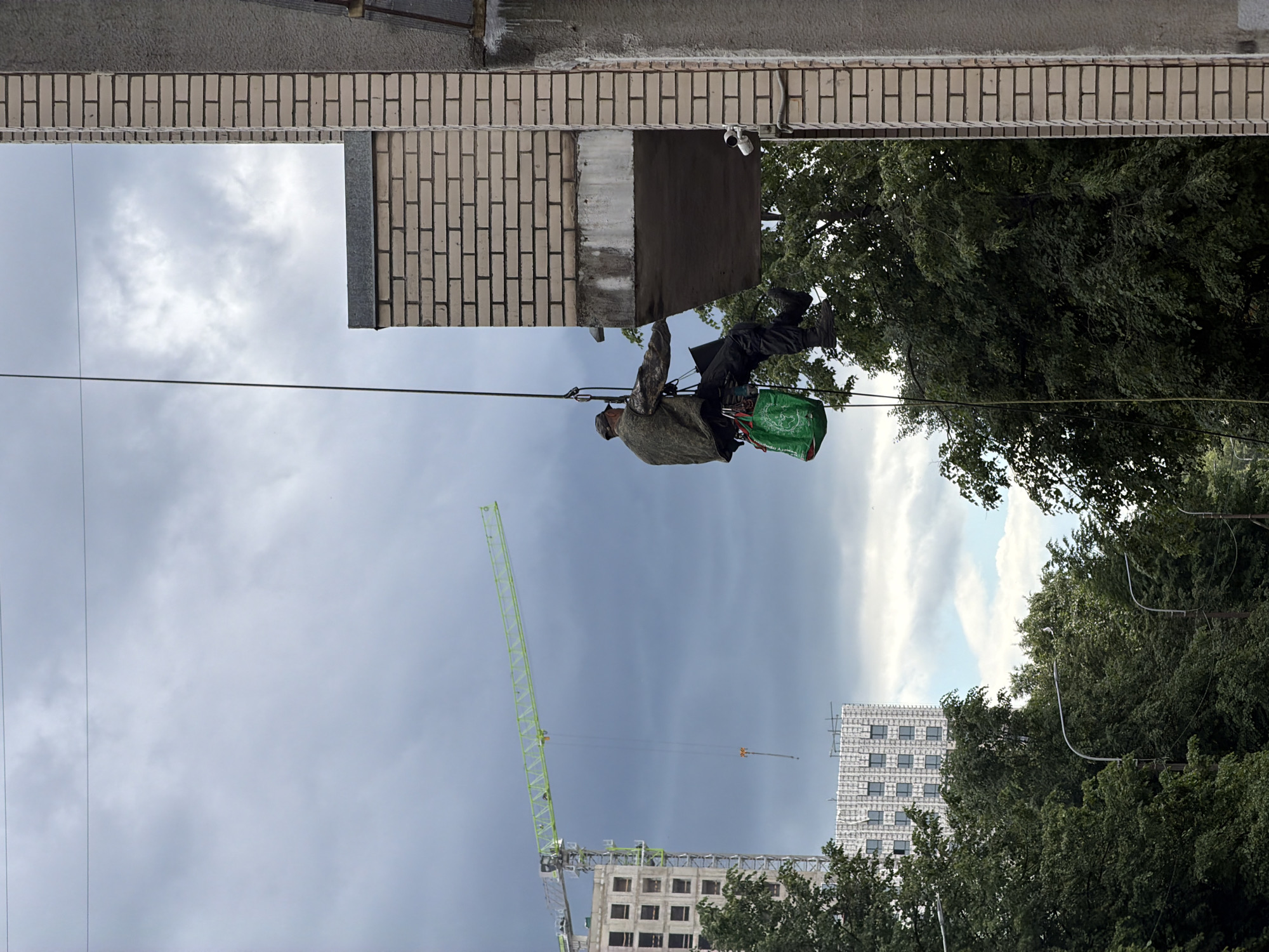 A man is rappelling down the side of a building in Saint Petersburg, Russia. He is wearing a camouflage jacket, dark pants, and a cap. He is secured by a rope and harness, and he has a green bag hanging from his side. The building is made of light-colored bricks and has a balcony. In the background, there is a construction crane and other buildings under construction. The sky is cloudy and overcast.