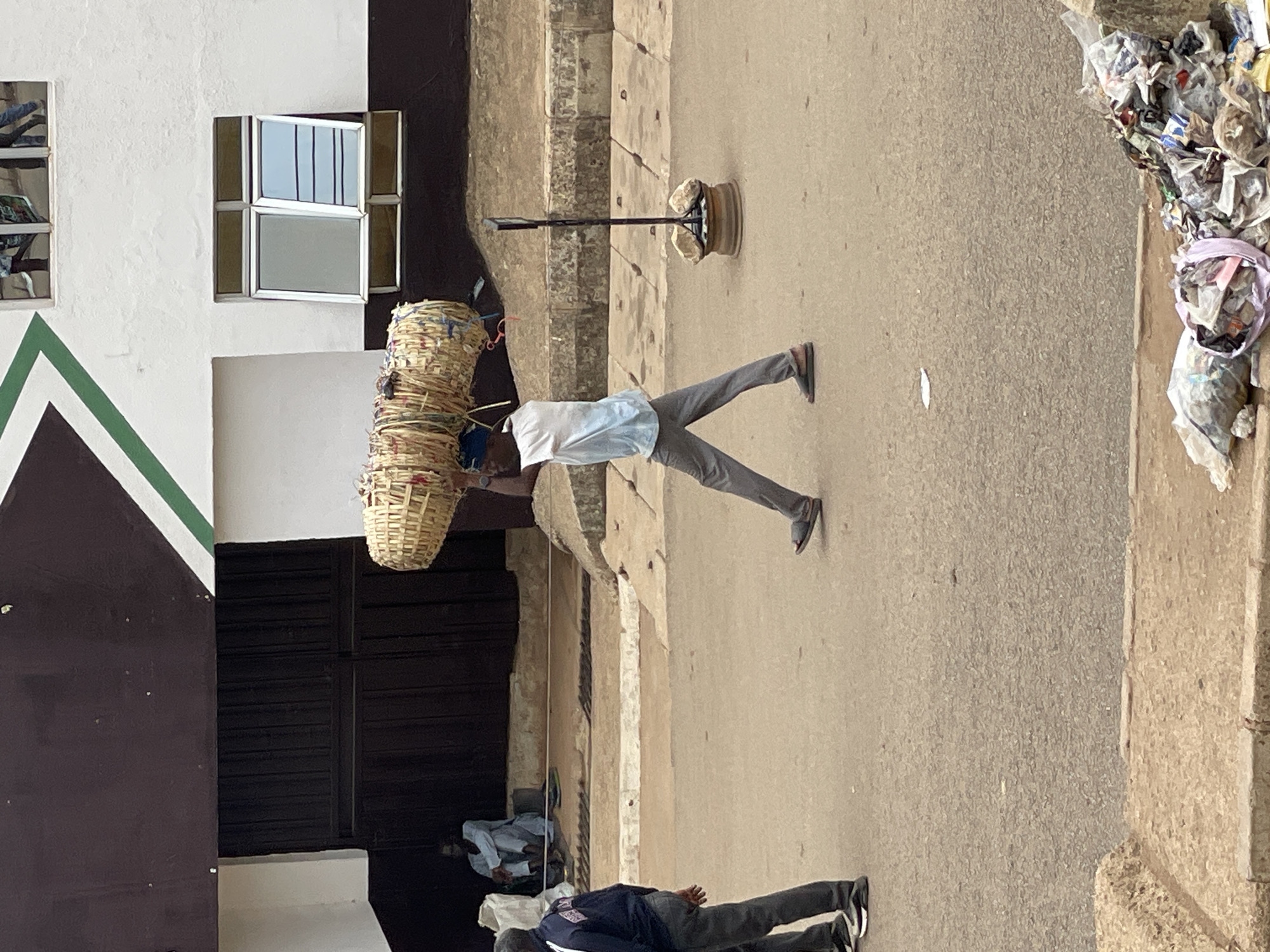 The image depicts a street scene in Bukuru, Nigeria. A man is walking in the middle of the street with four woven baskets stacked on his head. He is wearing a light blue shirt and gray pants. In the foreground, two people are lying on the ground; one is wearing a navy shirt and gray pants, and the other is wearing a light blue shirt. The buildings lining the street are a mix of white, brown, and black paint. One building has a white wall with two windows, one with a green trim. There is also a light pole with a sign, and a pile of trash in the upper right corner of the image. The street is made of a light gray material and appears to be paved.