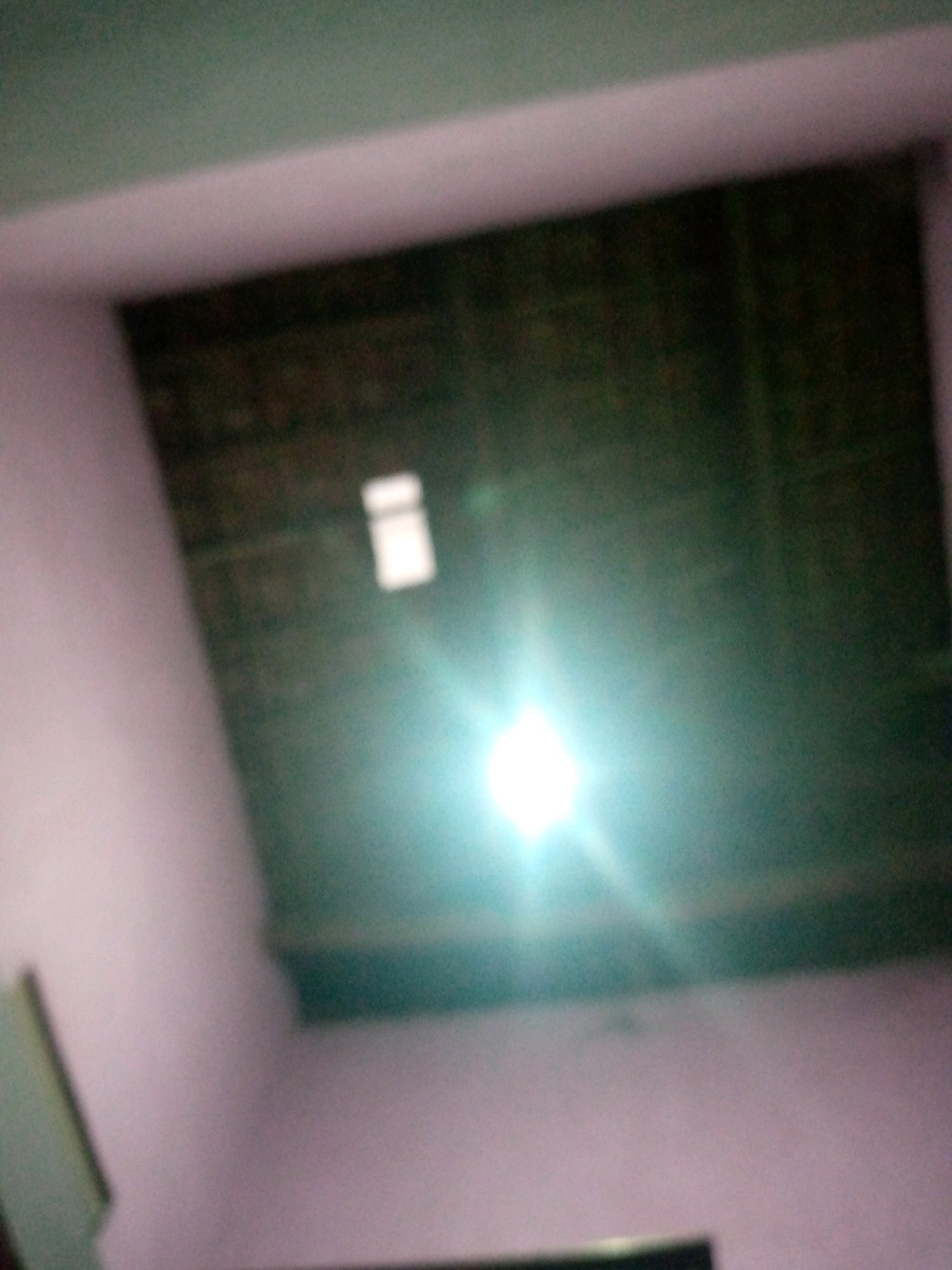 The image displays a poorly lit interior space, possibly a room, with a dominant bright light source causing significant glare and obscuring details. The walls appear to be painted a light purple or pink hue, with a darker section visible on the ceiling or upper wall. A rectangular opening, possibly a window or vent, is faintly discernible within the darker area. The overall impression is one of low light conditions and a slightly disorienting perspective, likely due to the intense light. The image does not contain any discernible elements to suggest a specific location within Sumberwono, Indonesia.