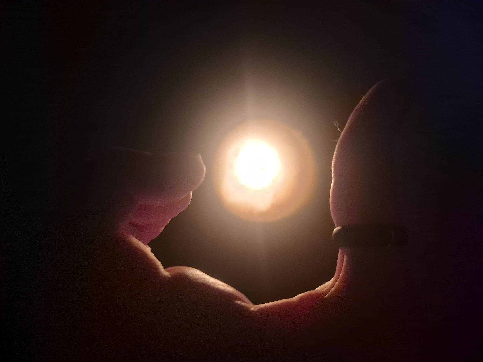 A close-up shot captures two human hands silhouetted against a singular, intensely bright, warm light source in an otherwise dark environment. The hands are positioned to form a heart shape, with the radiant light glowing centrally within the frame created by the fingers and palms.
The light emanates a soft, golden-orange hue, resembling a concentrated flame, an incandescent bulb, or a small lamp, with a strong lens flare effect around its core. Its brilliance contrasts sharply with the deep black background, indicating the photograph was taken in darkness, likely at night or in a dimly lit room.
The skin tone of the hands is visible where the light catches their inner contours, showing a natural, warm flesh color. The right hand distinctively wears a dark, simple band-style ring on one of its fingers. The texture of the skin, including faint wrinkles and pores, is discernible on the parts illuminated by the light.
The deliberate framing of the light by the hands forming a heart shape evokes themes of tenderness, love, hope, or holding onto something precious. There is no visible text in the image. While the location context is Chaikovsky, Russia, no specific visual cues within the image itself confirm this.