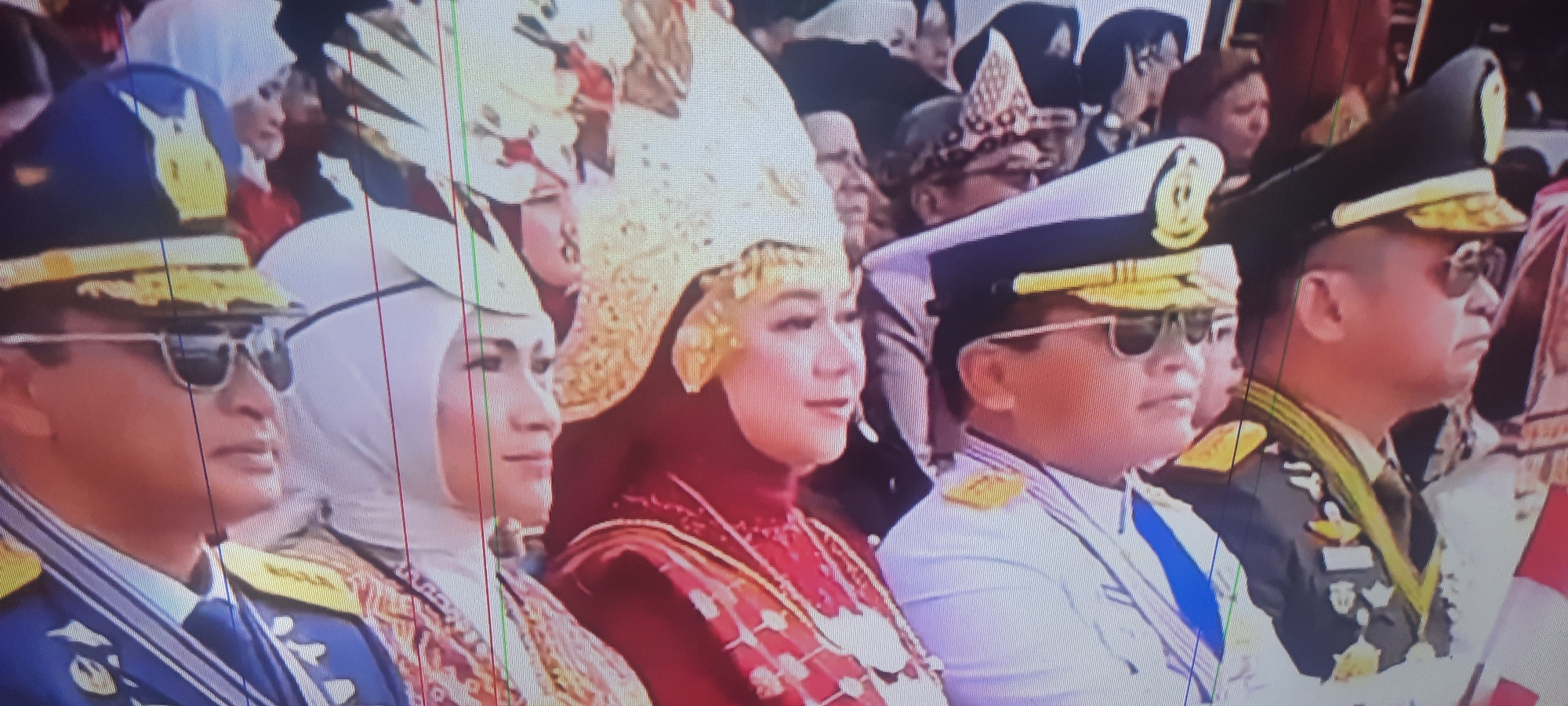 This is a medium shot, high-angle, slightly blurry, horizontal video still image of a group of people. In the foreground, from left to right, are three men and two women. The leftmost man wears a dark blue military-style hat with a gold brim and a yellow emblem, sunglasses, and a dark blue uniform with a white shirt and tie, and a sash across his chest. Next to him, to his right, is a woman wearing a white hijab and a colorful patterned garment. To her right, a woman wears a large, ornate, golden headdress and a red garment. Next to her is a man in a white uniform with a white cap and gold trim, and sunglasses. To his right, a man in a dark military uniform with a dark cap, gold trim, and a yellow and green sash is visible. Several other people are in the background. The image appears to be taken outdoors during the day. The scene depicts an event that could be a formal ceremony or procession. The text in the prompt states the location is Jatitengah, Indonesia, however there are no visual cues within the image to confirm or deny this information.