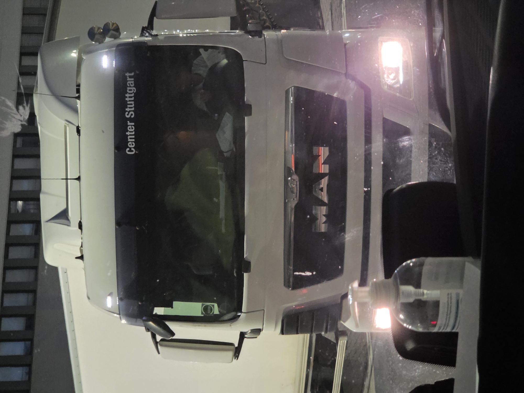 The image shows the front of a white MAN truck parked outdoors at night, possibly in Walldorf, Germany. The truck has a dark grille with the "MAN" logo prominently displayed. Above the grille, "Center Stuttgart" is written in white text on the dark windshield. The headlights of the truck are illuminated, casting a bright light. Inside the cab, two figures are visible, one wearing a light-colored shirt and the other appearing to be reading something. A bottle of hand sanitizer is placed on the dashboard in the foreground, with a red light reflecting off its surface. In the background, a multi-story building with dark windows is visible. The overall scene suggests the truck is parked in an industrial or commercial area.