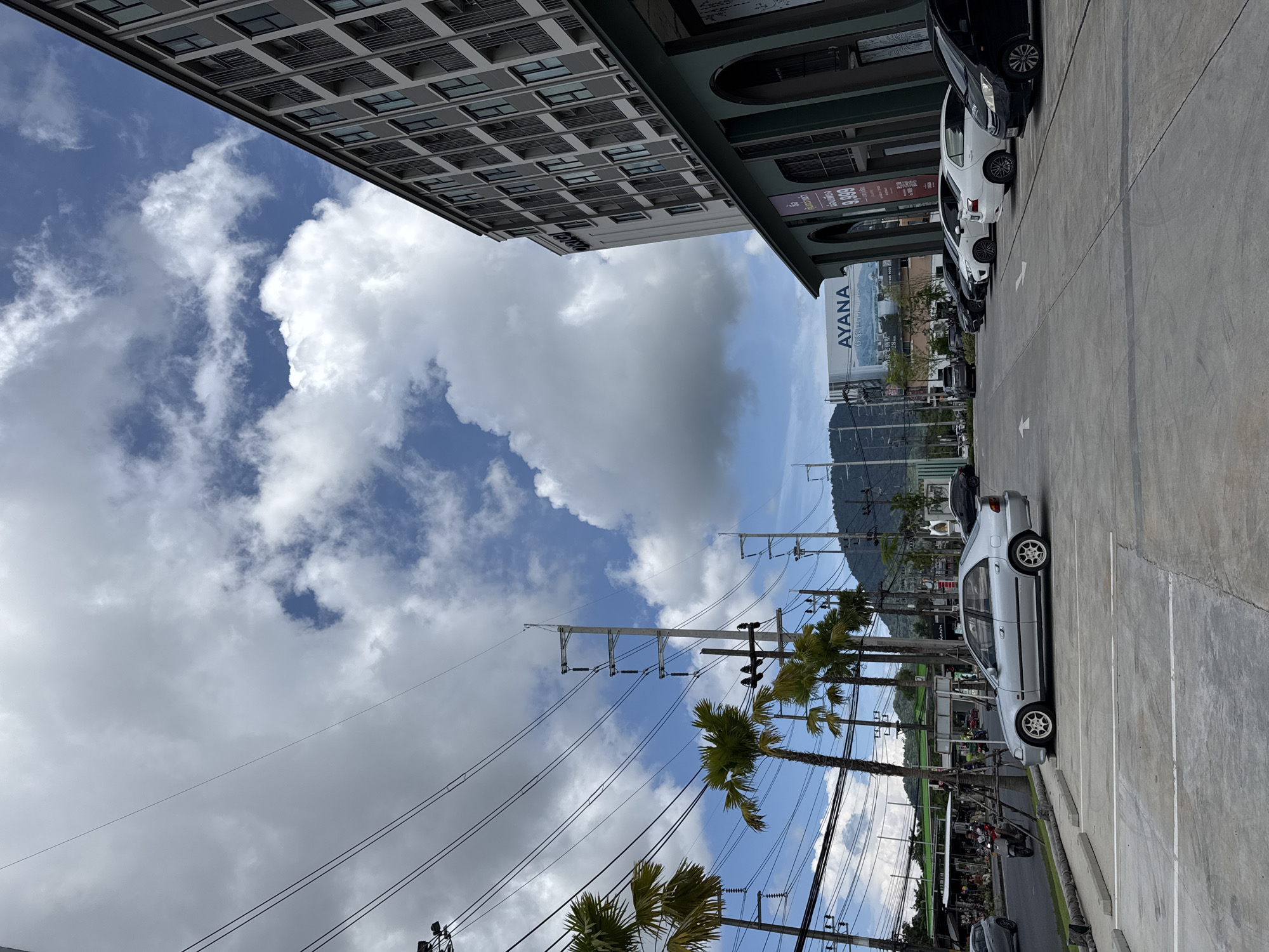 A long shot captures a parking lot in front of a modern building in Kathu, Thailand. The building, with its grey facade and rows of windows, dominates the right side of the frame. To the left, palm trees line a street with a blue sky dotted with white clouds overhead. Several cars are parked in the lot, and a motorcycle is visible on the street. Power lines crisscross the sky, and a billboard for "AYANA" is present in the background. The scene appears to be during the daytime.