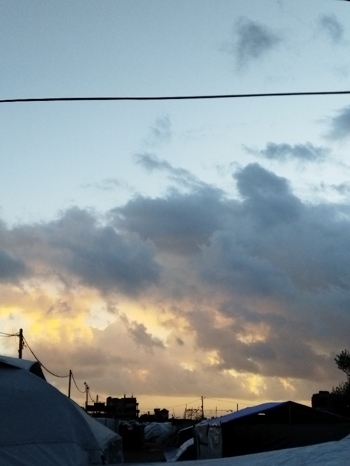 An outdoor scene in Khan Yunis, Palestinian Territory, depicts a twilight sky with a transition from light blue at the zenith to heavy, dark grey clouds interspersed with areas of golden-orange light near the horizon. A prominent black utility line spans horizontally across the upper frame. In the lower third, the silhouetted shapes of numerous structures are visible. On the left, a large, rounded, light-colored tent or temporary shelter occupies the foreground. To its right, several other lower-profile shelters, covered in light-toned tarpaulins, are arranged. Utility poles with visible wires punctuate the mid-ground. In the distant background, dark silhouettes of multi-story buildings are observable against the lighter sky. No individuals or specific actions are discernible. The overall environment suggests an urban or peri-urban setting featuring temporary housing under a dramatic sky.