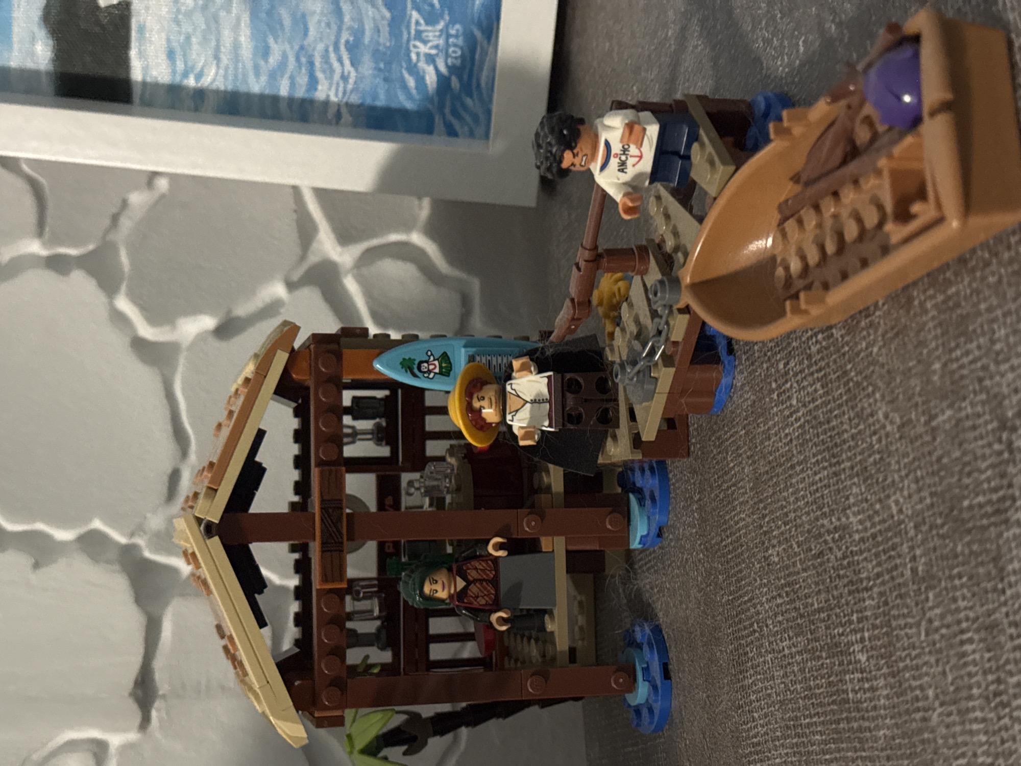 The image displays a Lego setup featuring minifigures and structures, likely depicting an island or tropical scene, resting on a grey, textured surface.
Three Lego minifigures are present. One, with black curly hair and light skin, wears a white shirt with the word "ANCHO" and an anchor logo, and is positioned near a small, light brown Lego boat. This boat contains various brown Lego elements and a round purple piece. Another minifigure, also light-skinned, wears a yellow hat and a white and dark-colored shirt, standing next to a blue surfboard adorned with a hula dancer graphic. The third minifigure, with green hair and a patterned dark top, is situated inside a brown Lego hut or shack.
The hut is constructed from brown Lego bricks with light tan roof sections. It rests on blue circular bases, suggesting a floating or stilt-like structure, and features chain or net-like details. Behind the Lego scene, a framed picture is visible, displaying abstract blue and white art, possibly representing water. Partially visible text on this picture includes "Kat" and "2025". The overall lighting suggests an indoor setting.