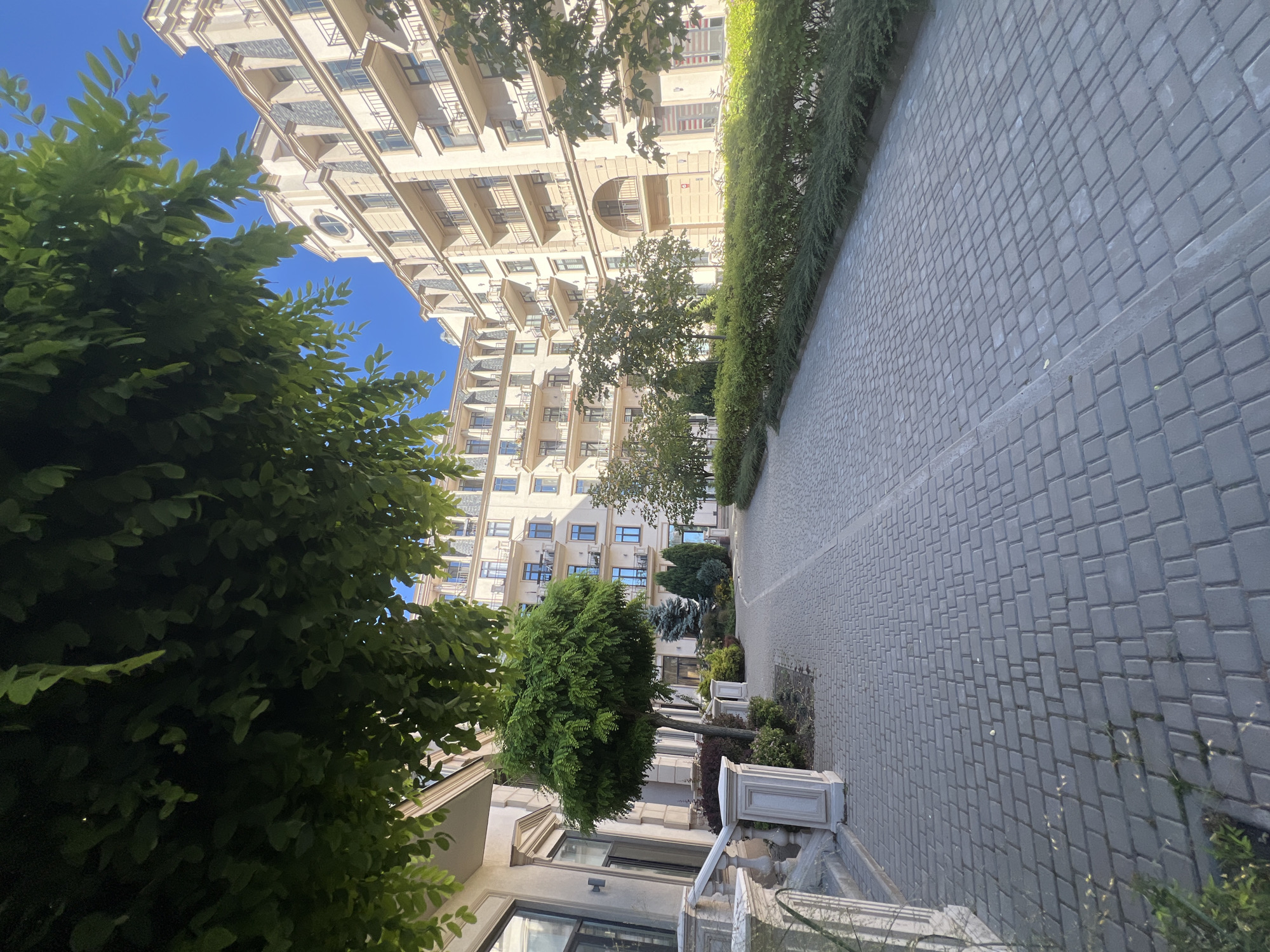 The image depicts a perspective view of a paved walkway leading towards a modern building complex in Fontanka, Ukraine. The walkway is constructed from grey rectangular pavers arranged in a herringbone pattern. To the left of the walkway, a building with beige facade and multiple windows is visible, with ornate white railings leading up a set of stairs. A well-maintained, rounded tree stands prominently on the left side of the walkway, surrounded by low green shrubs. Further down the walkway, more trees and meticulously trimmed hedges line the path. The building complex itself is multi-story, with a beige exterior, numerous balconies, and dark-tiled roofs. The sky above is clear and blue. The scene is bathed in natural daylight, suggesting it was taken during the daytime. No people are present in the image.