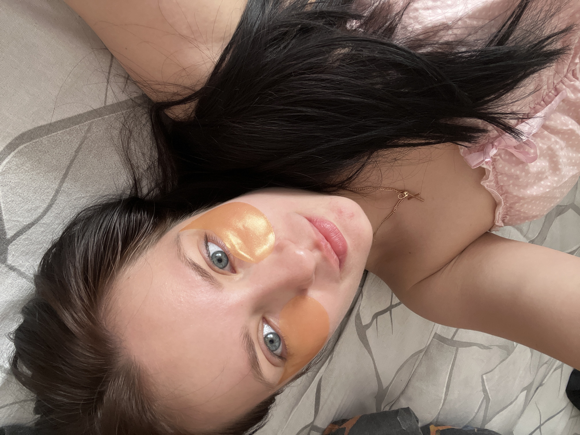A woman lies on a bed covered with a gray and white patterned sheet. She is wearing gold-colored eye patches under each eye and a pink and white polka dot top with a ruffle detail. Her dark hair is spread across the bed. She is wearing a delicate gold necklace with a small charm. The location cannot be determined from the image.