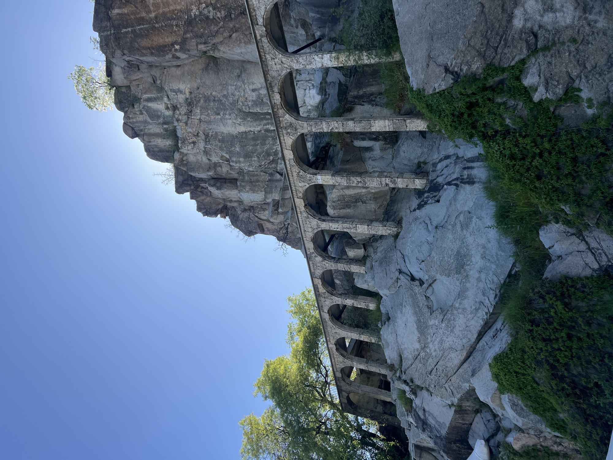 The image depicts an ancient Roman aqueduct in Plovdiv, Bulgaria, built on a rocky cliff face. The aqueduct consists of multiple stone arches. Green foliage grows on and around the rocks. The background is a clear blue sky. The aqueduct is situated on a rugged, uneven terrain. The sun is shining brightly, and a tree grows from the cliff at the top left.