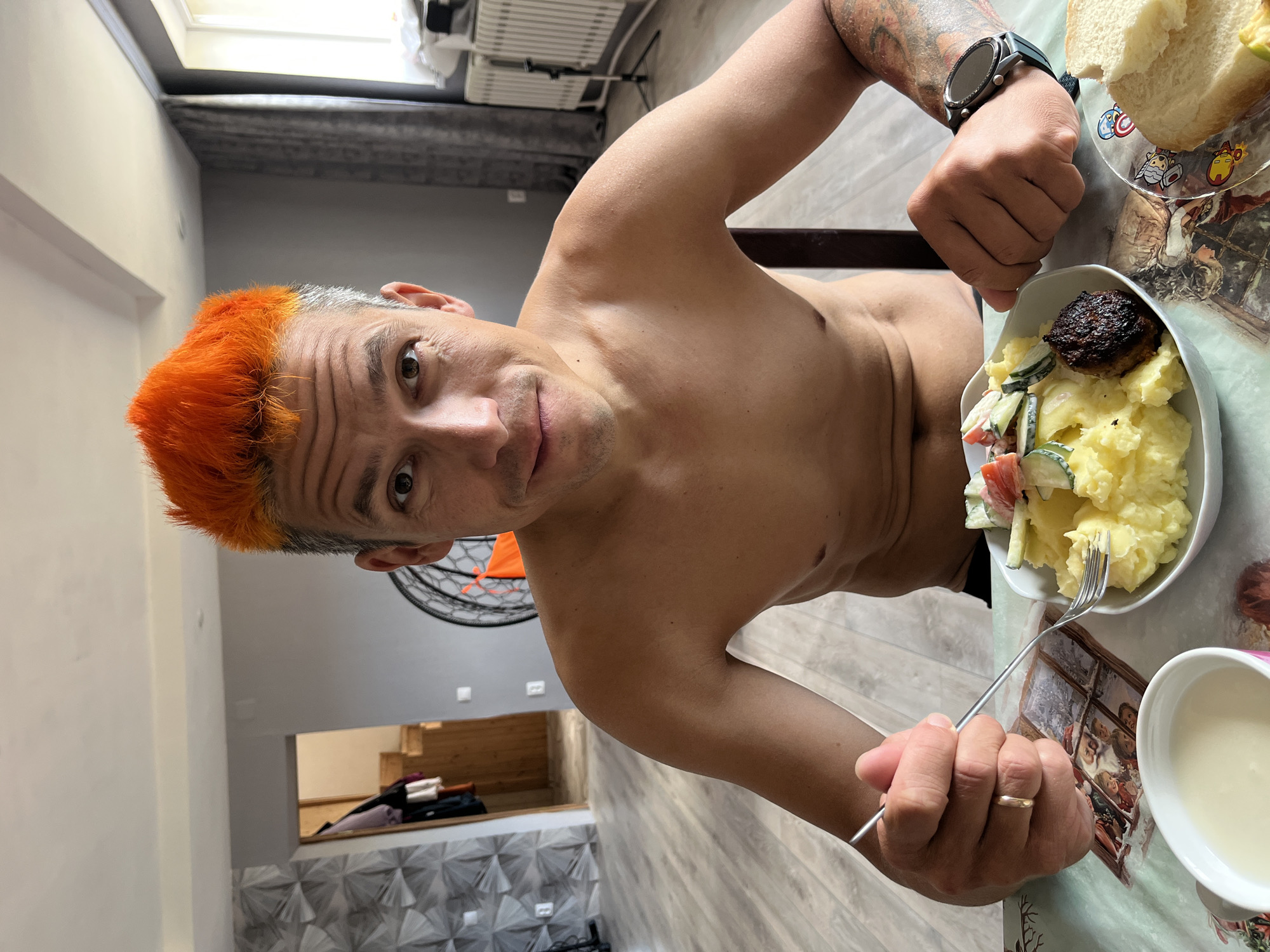 A shirtless man with bright orange hair is seated at a table, holding a fork in his right hand and looking at the camera. He has a wedding ring on his finger. On the table, there is a plate with mashed potatoes, a salad, and a meat patty, a bowl of liquid, and a plate with bread. The table has a Christmas-themed tablecloth. A smartwatch is visible on his left wrist. In the background, there is a room with light grey walls, a window with curtains, and an open doorway leading to another room. A radiator is visible near the window. A piece of exercise equipment is behind the man. The image was taken in an apartment in Chaikovsky, Russia.