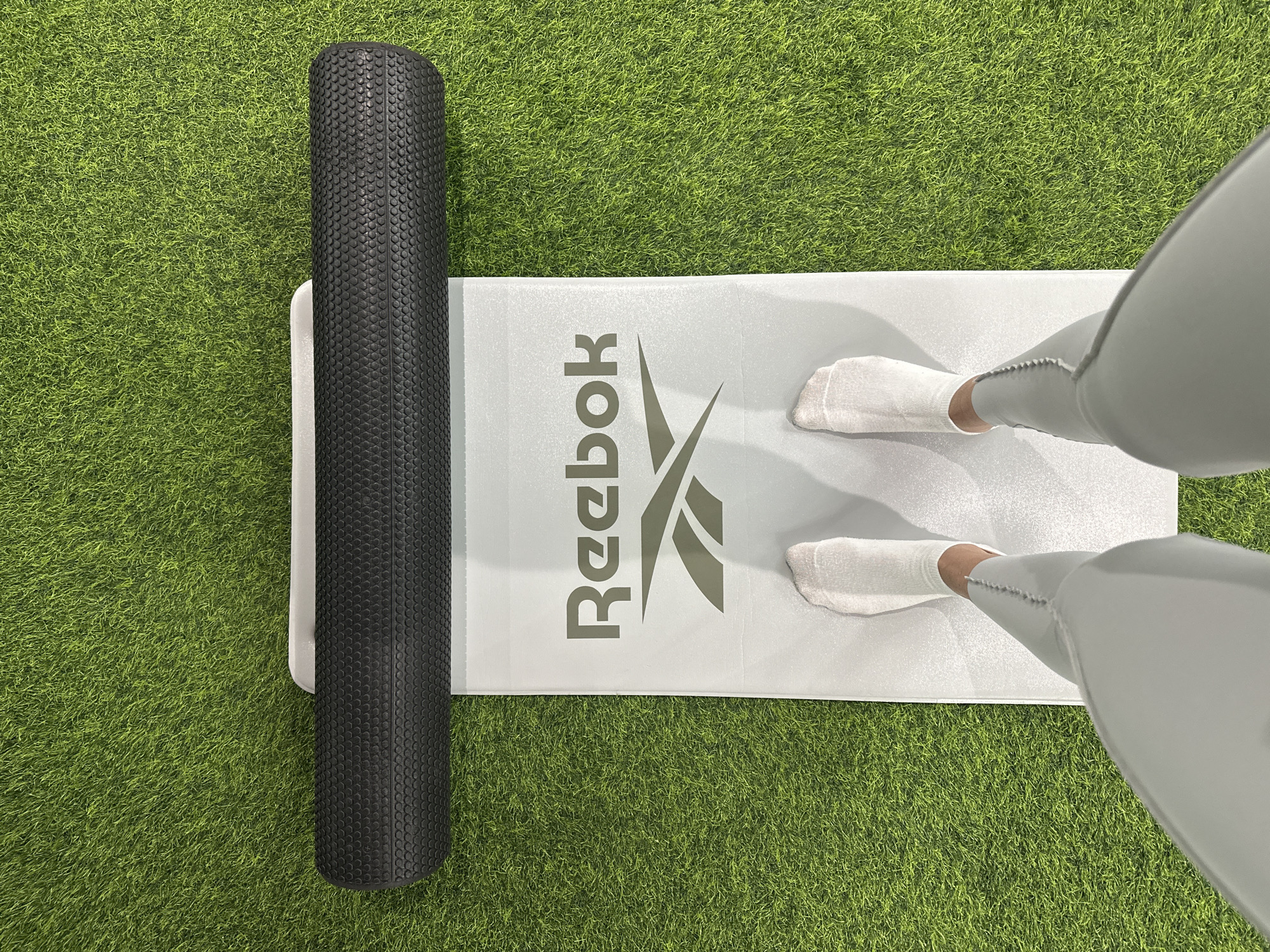 A pair of feet wearing white socks and light gray leggings stands on a white yoga mat. The mat has a dark gray Reebok logo on it. To the left of the mat, a black foam roller lies on a green, artificial turf surface. The image is taken from a top-down perspective. The lighting suggests an indoor setting or an overcast day. There is no specific indication of time of day, weather, or emotion. The location context provided is Irkutsk, Russia, but there are no visual cues in the image to confirm this.