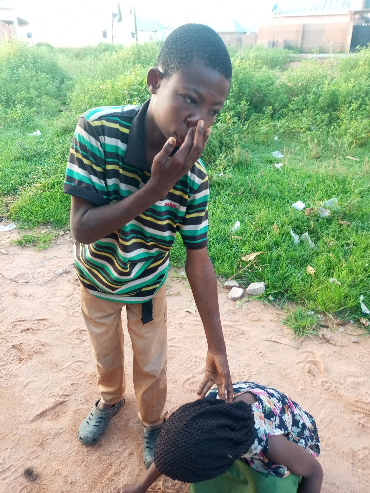 In the city of Tempure, Nigeria, a young boy wearing a striped t-shirt and tan pants stands over a person lying on the ground. The person on the ground is wearing a floral dress and a black head covering. The boy is touching the face of the person on the ground. The scene appears to be outdoors in a sandy area with some vegetation and scattered debris.