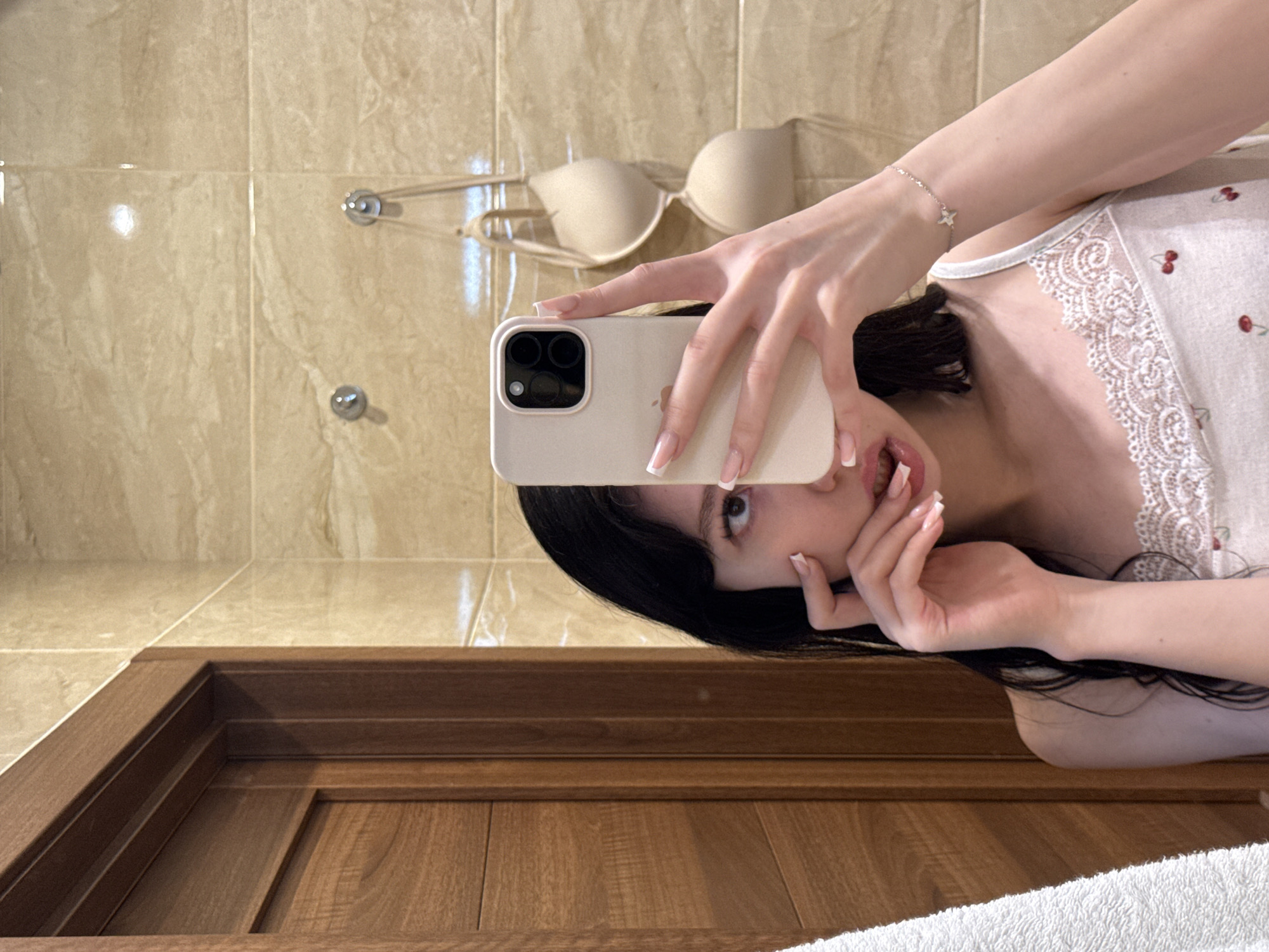 A young woman with dark hair takes a mirror selfie in a bathroom. She is wearing a white tank top with lace trim and cherry embroidery. A beige bra hangs on the wall behind her. The bathroom has beige tiled walls and a wooden doorframe. A white towel is visible on the left side of the frame. The woman is holding a white smartphone to take the picture, and her left hand is touching her face. The scene appears to be in a residential bathroom.